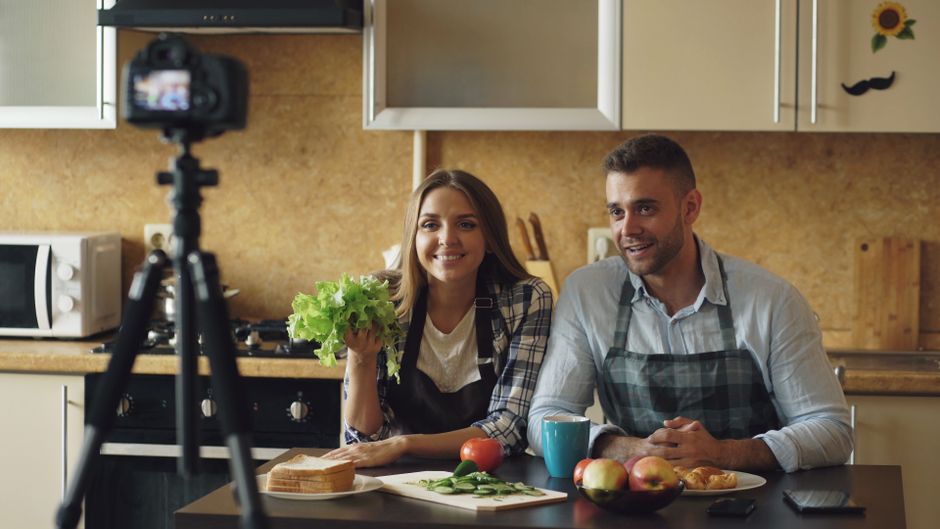 Couple recording a cooking video in their kitchen with fresh vegetables and fruits, smiling at the camera.