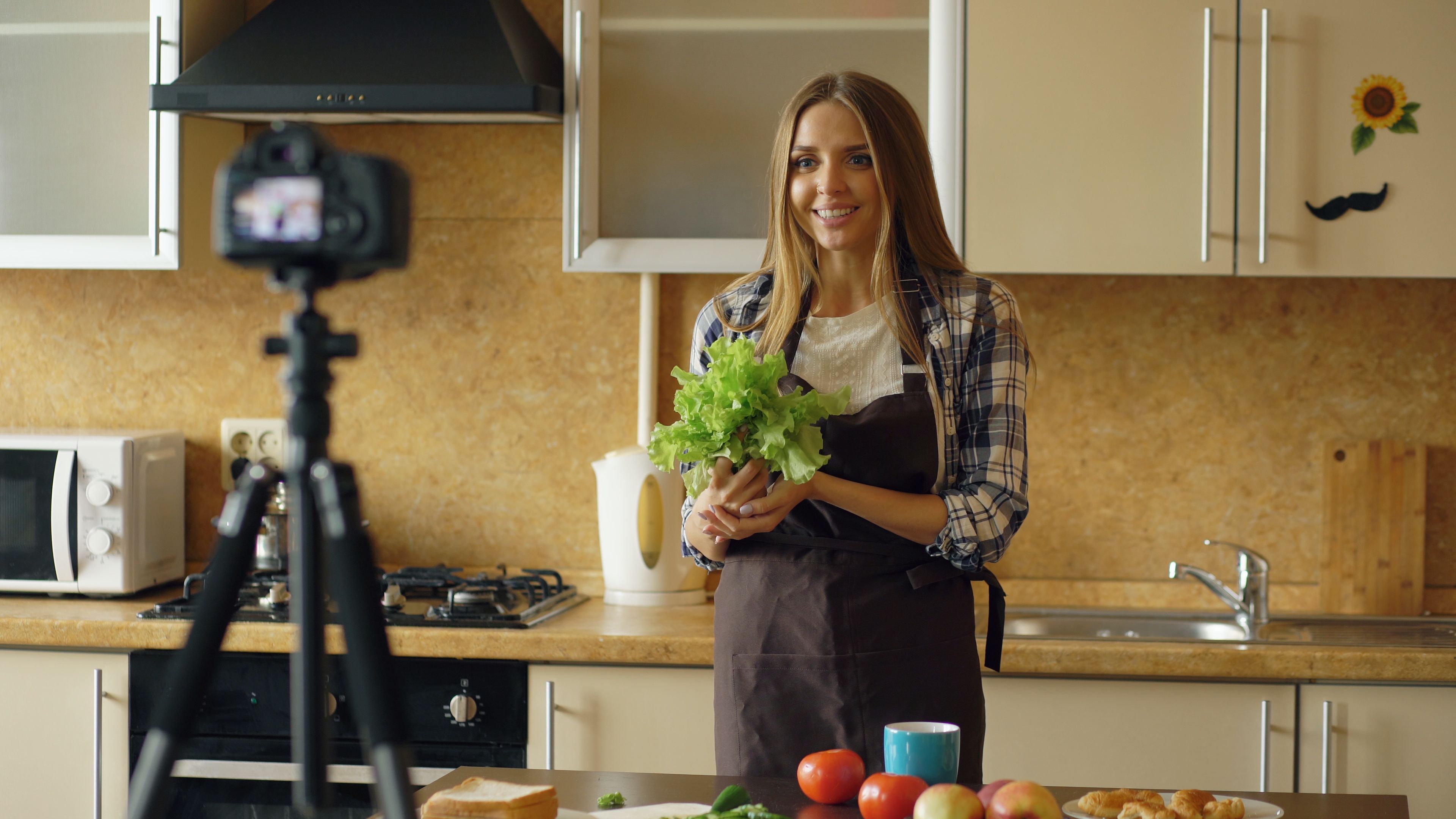 Woman filming a cooking video in a kitchen, holding fresh lettuce. Ideal for food and lifestyle content.