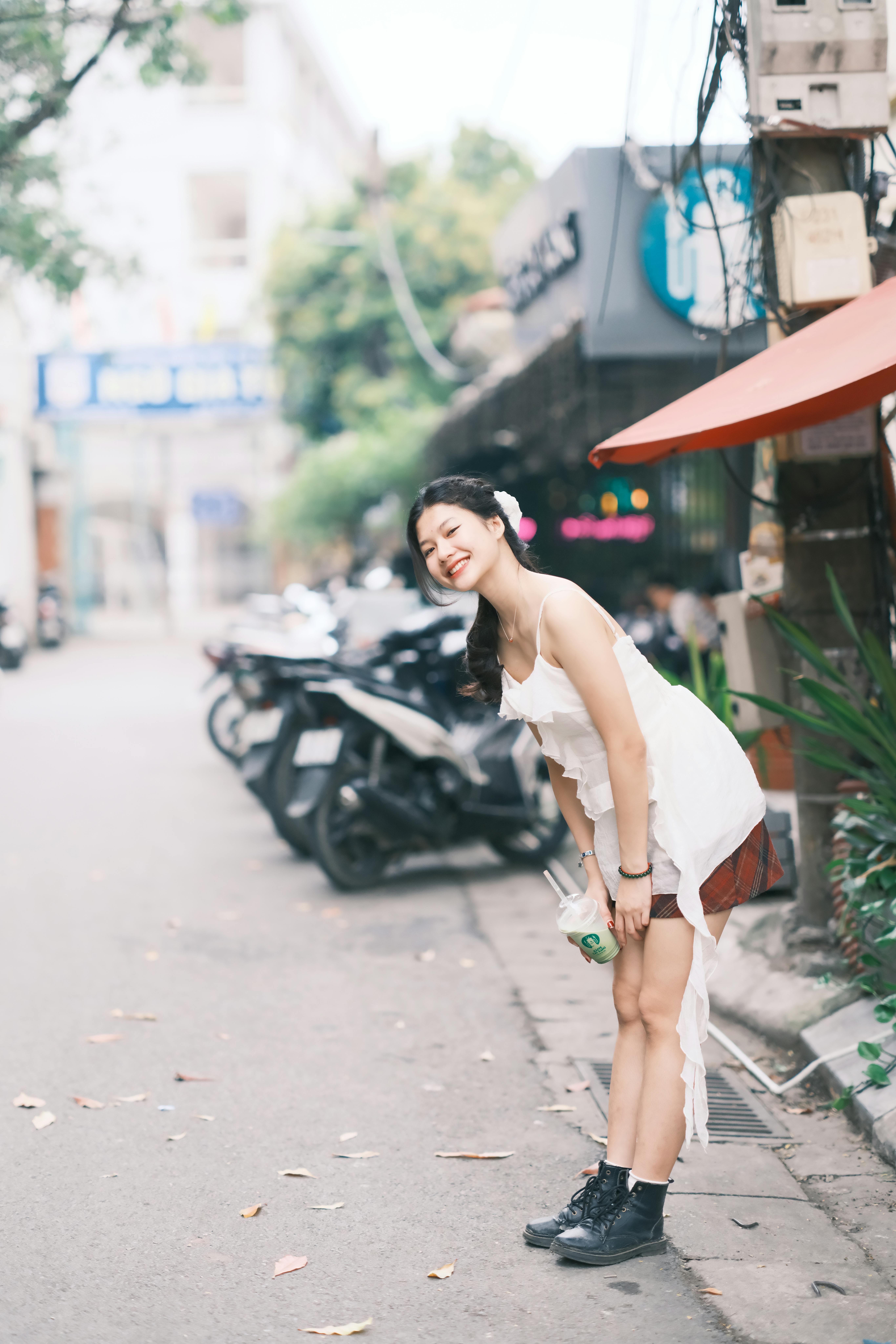 A joyful young woman in a white dress smiling brightly while standing on a lively street in Vietnam.