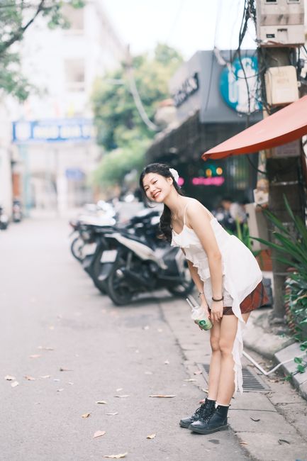 A joyful young woman in a white dress smiling brightly while standing on a lively street in Vietnam.