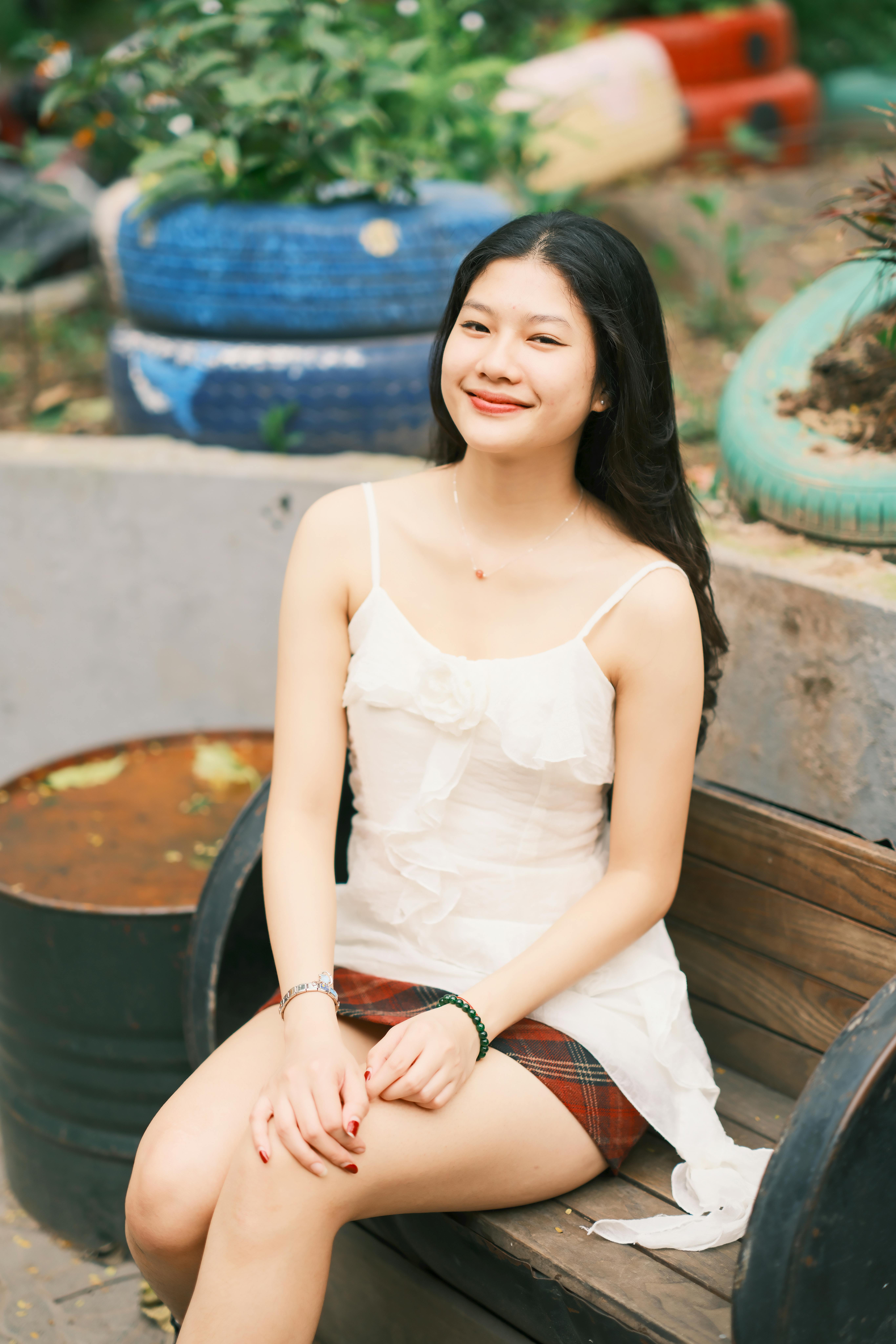 Portrait of a young Asian woman sitting on a bench in an outdoor garden, smiling warmly.