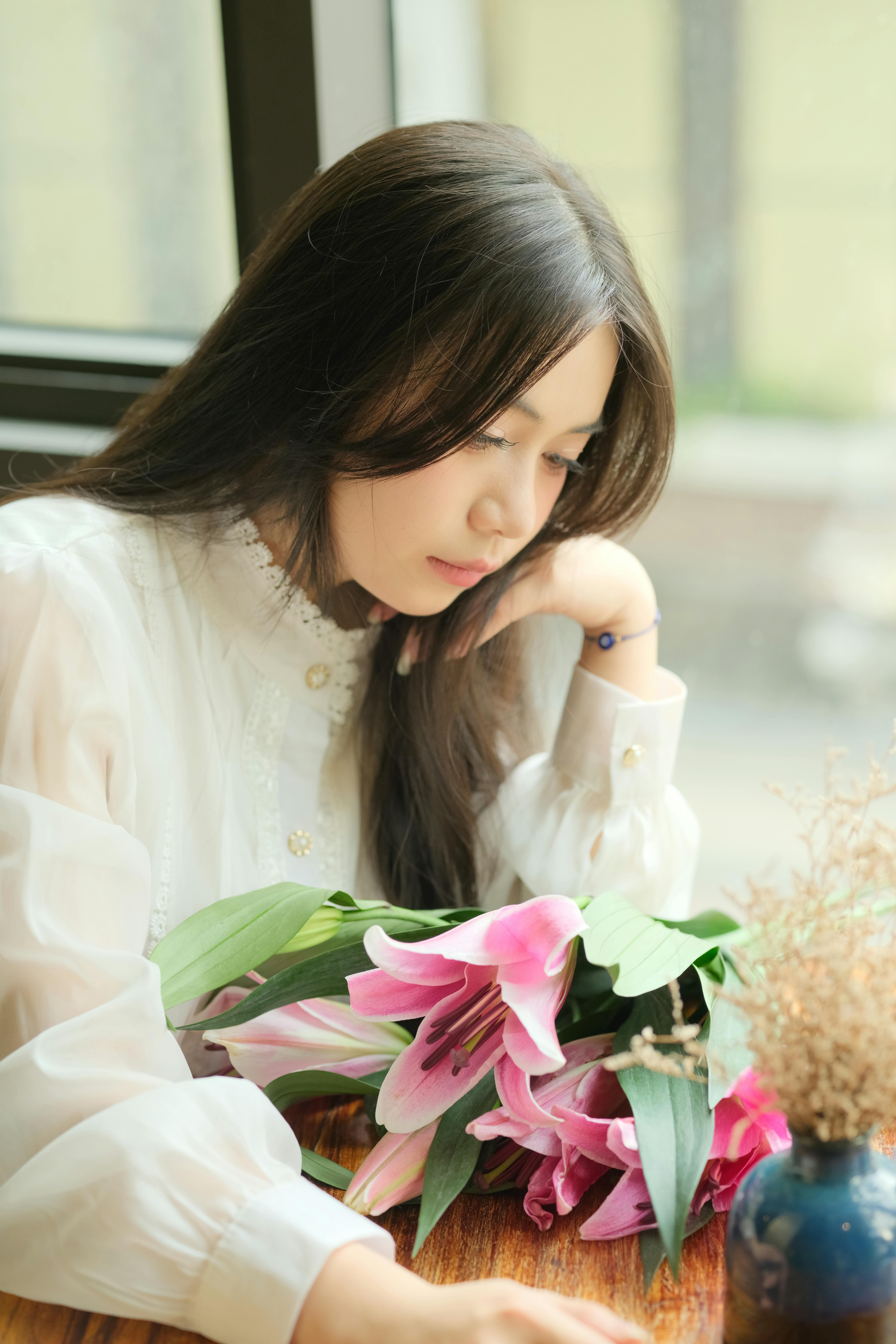 A contemplative woman with pink lilies sits indoors in Vietnam, captured in soft lighting.
