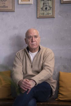 一位年长的男子坐在室内，背景是镶框的墙壁装饰，表情若有所思