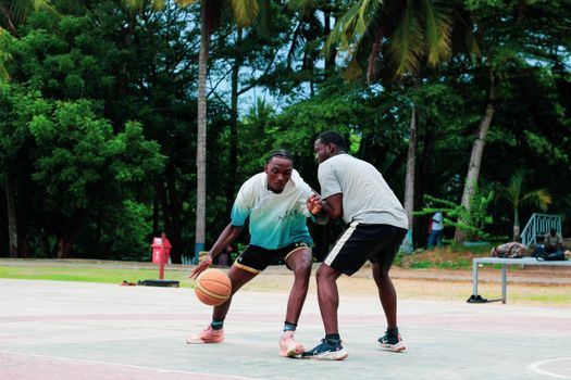 Two young men engaging in a competitive basketball game on an outdoor court surrounded by trees.