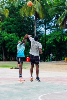 Two young men actively playing basketball in an outdoor court surrounded by lush green trees.