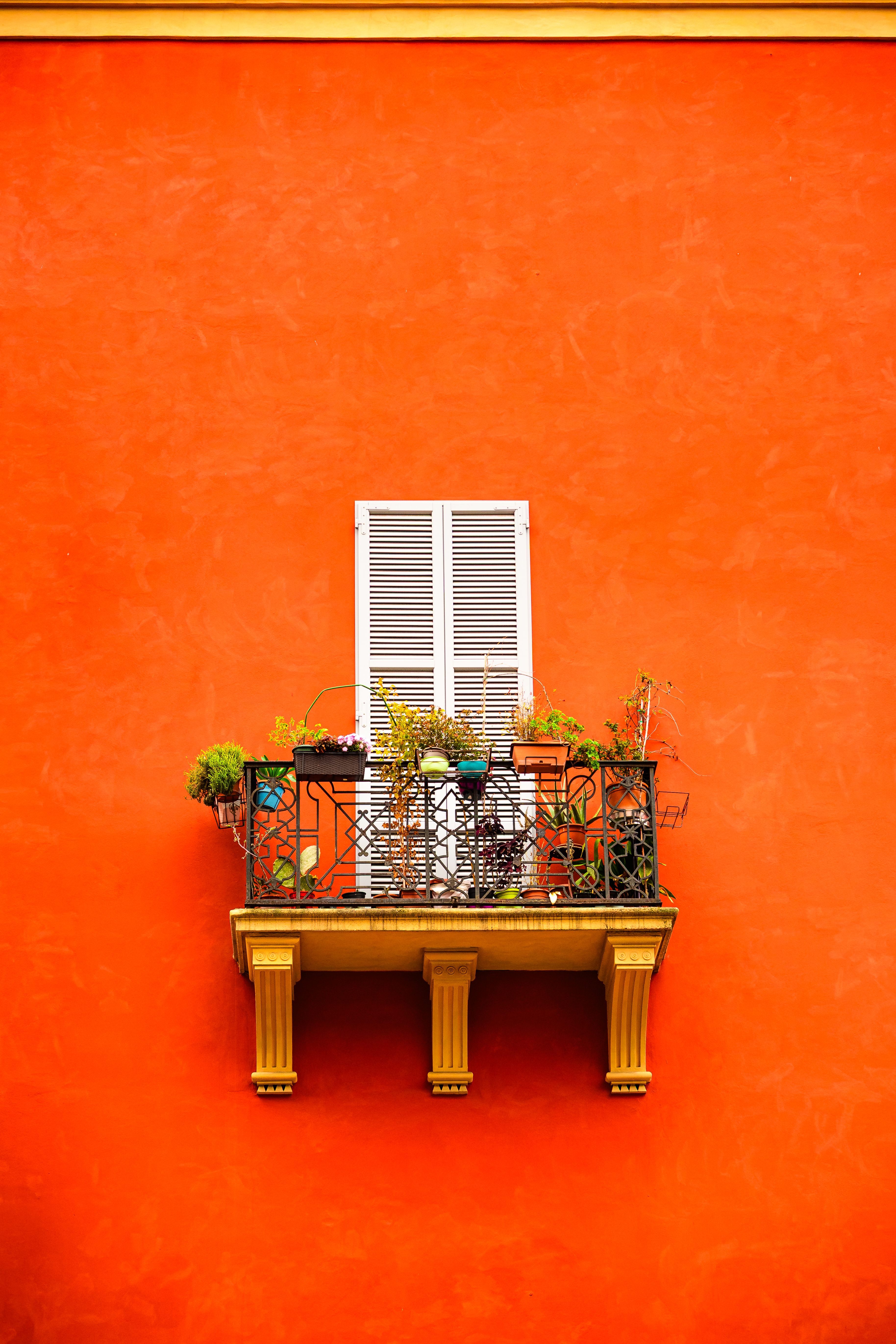 Orange wall with a decorative balcony and white shutters, showcasing colorful plants.