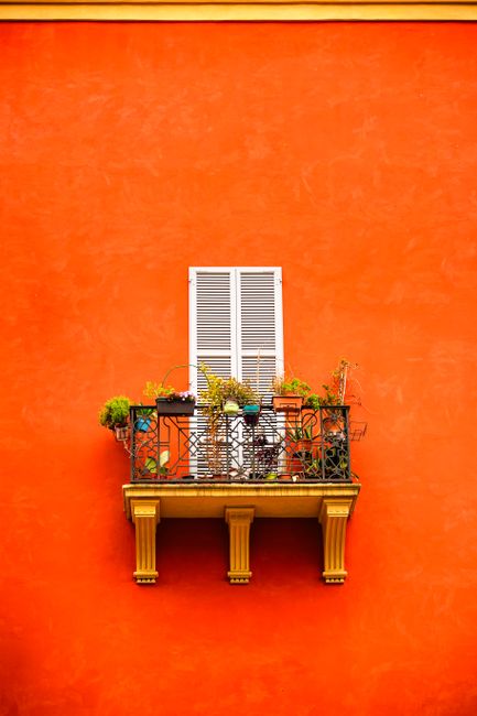 Orange wall with a decorative balcony and white shutters, showcasing colorful plants.