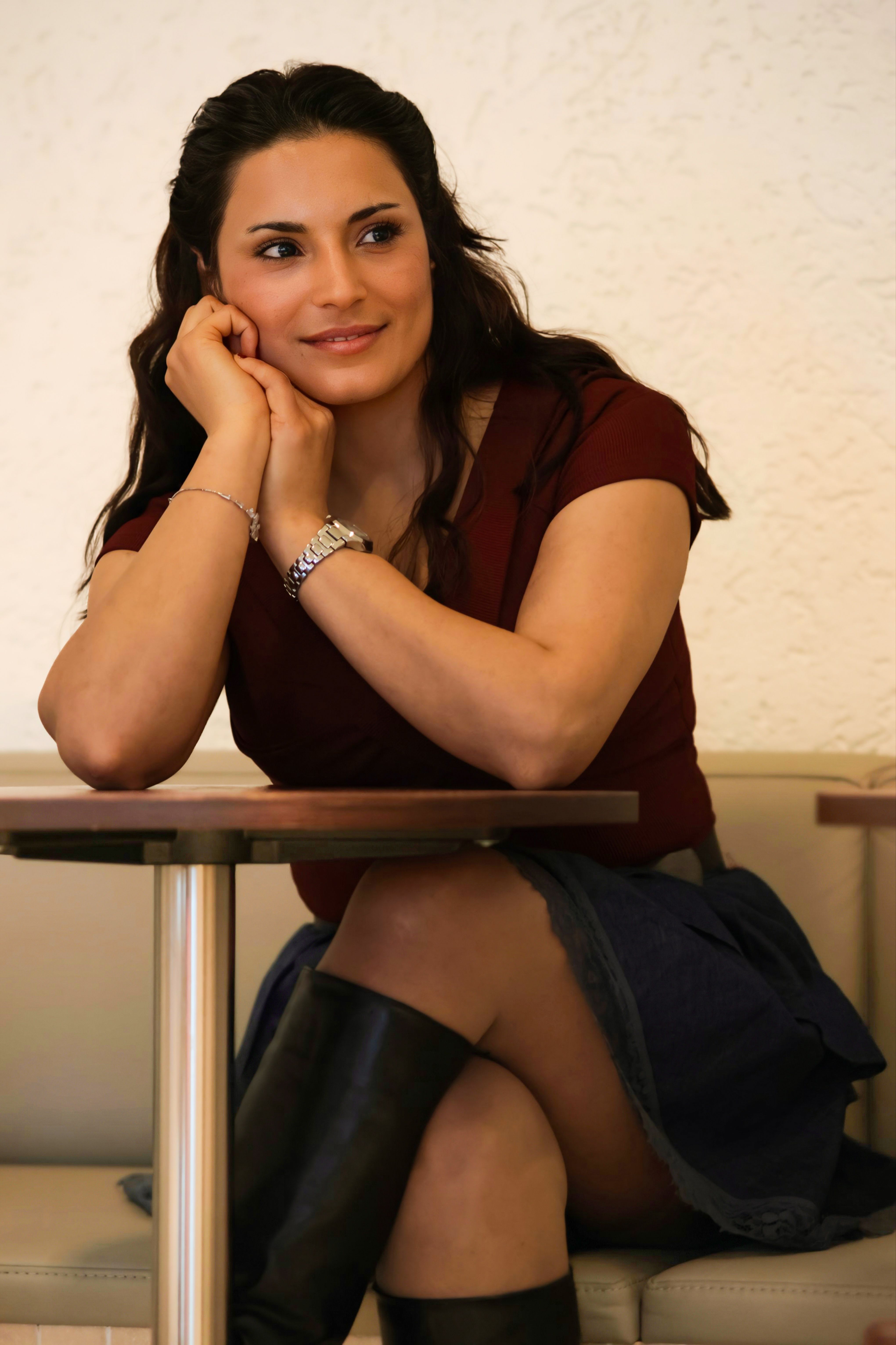 A woman with long brown hair smiles while sitting at a cafe table, wearing a maroon top.