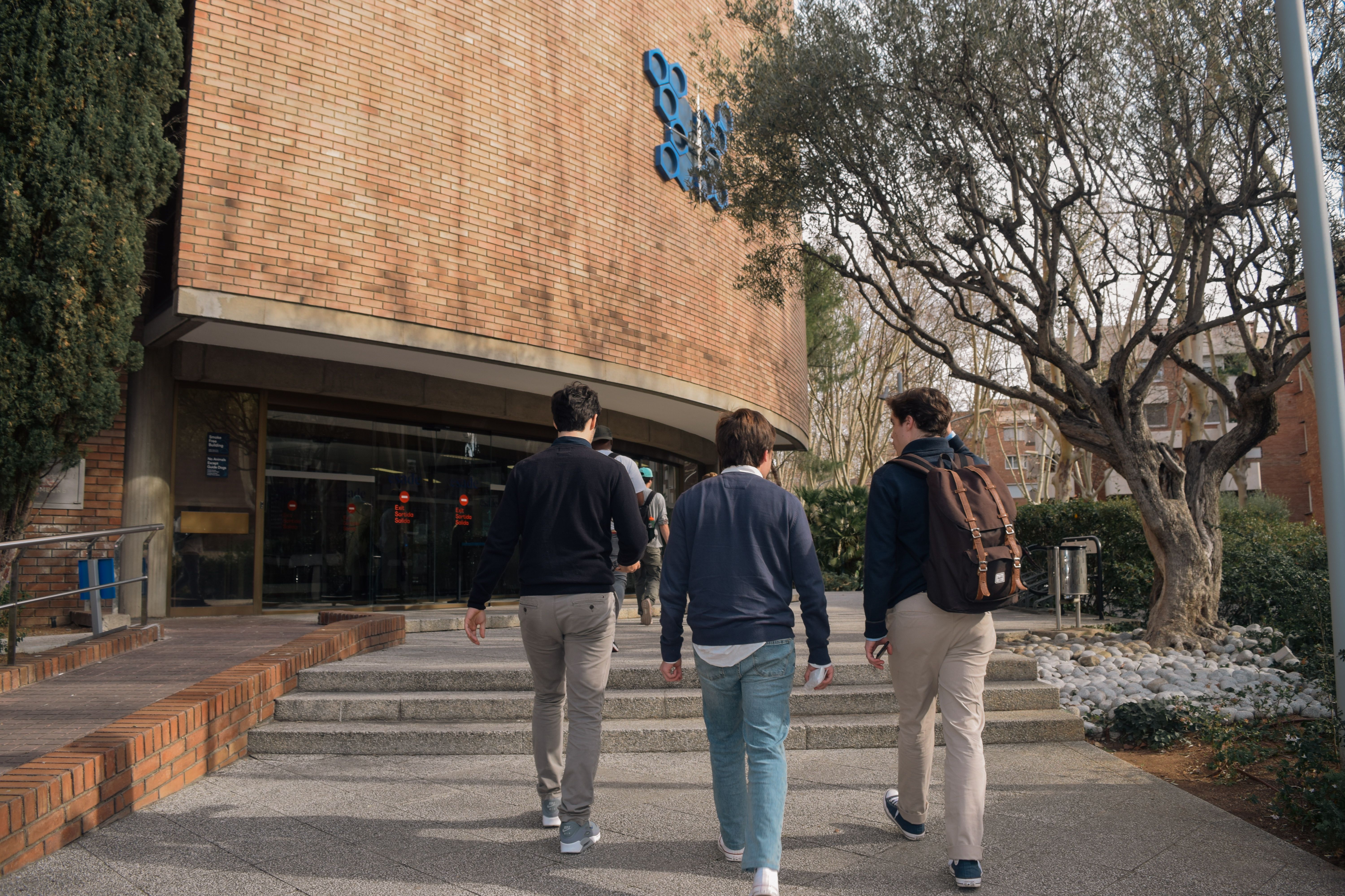 Three students approach a university building entrance in Barcelona, Spain on a sunny day.