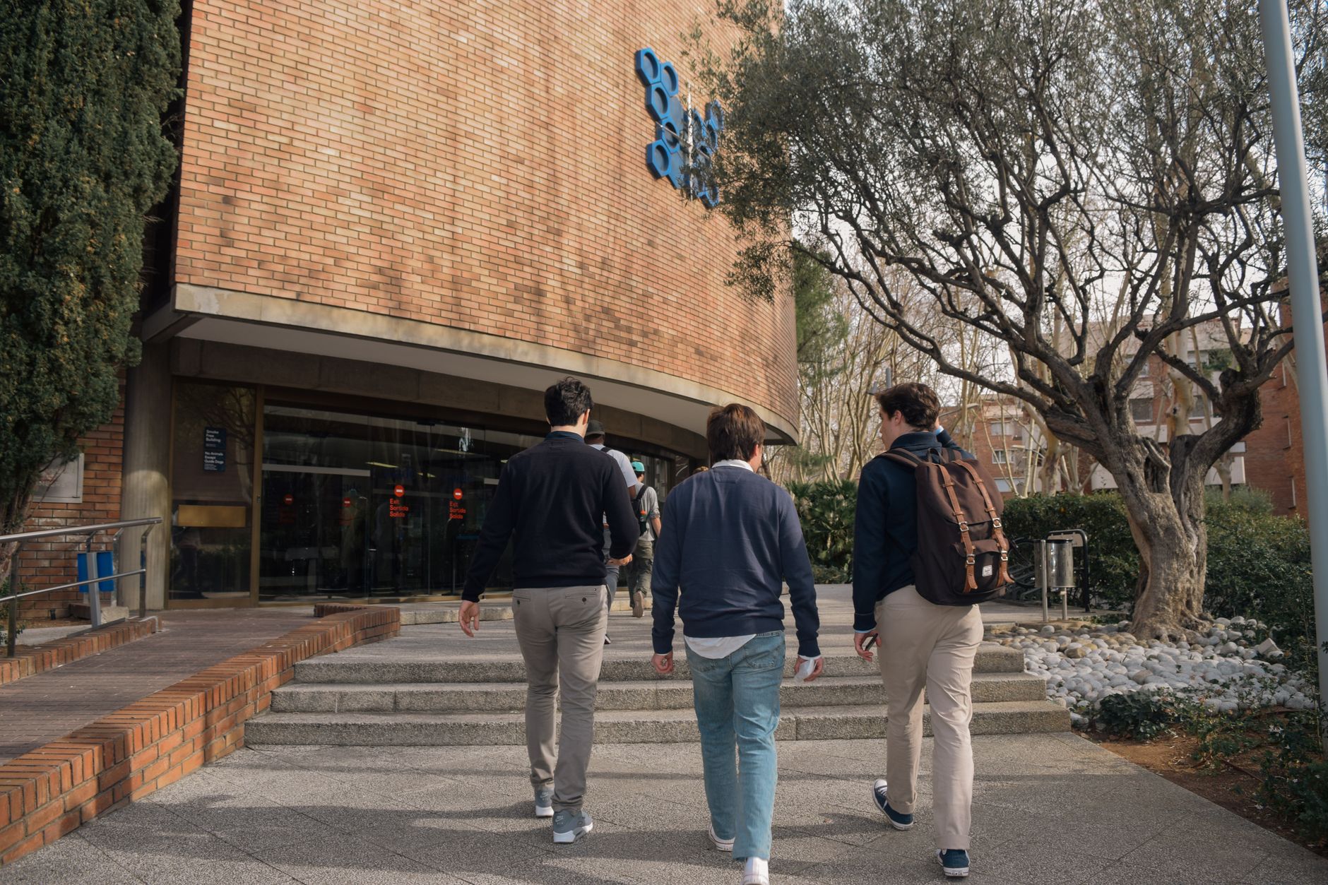 Three students approach a university building entrance in Barcelona, Spain on a sunny day.