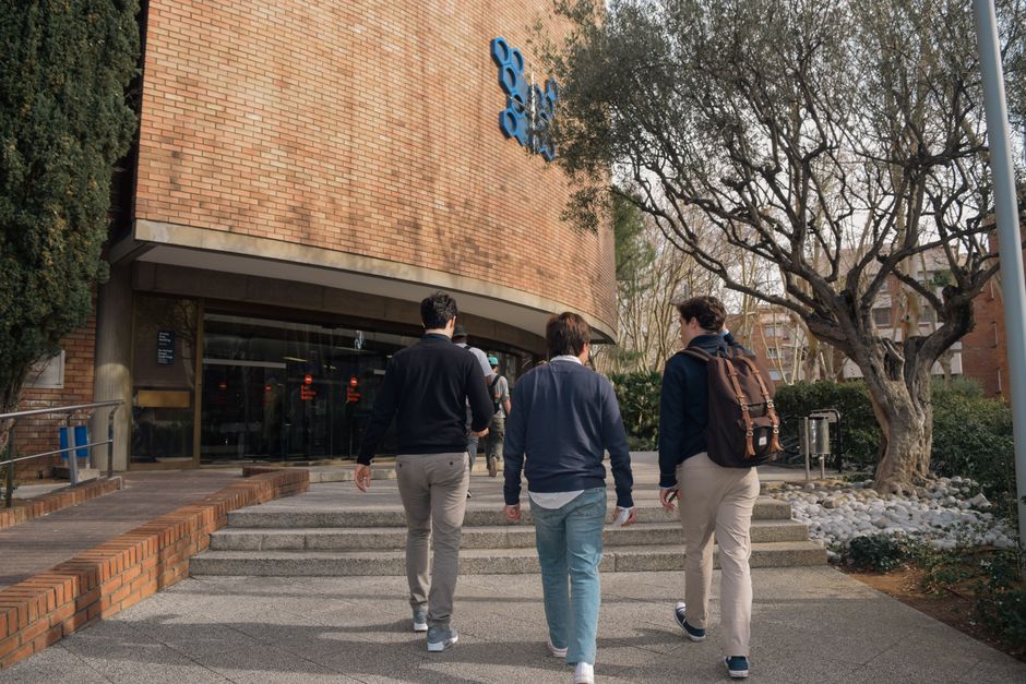 Tres estudiantes se acercan a la entrada de un edificio universitario en Barcelona, España, en un día soleado.