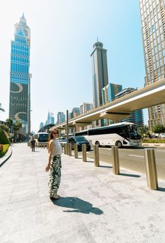 Woman in sunglasses and hat walks by Dubai skyscrapers under bright daylight.