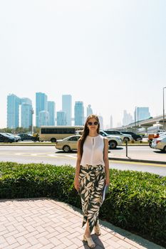 Stylish woman in sunglasses walks through Dubai with skyscrapers in the background.