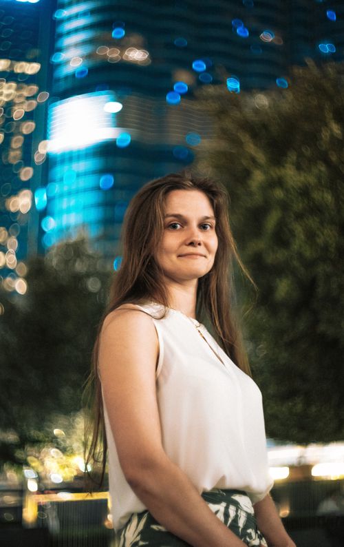 Free A woman stands confidently with the blurred lights of Dubai's skyline behind her at night. Stock Photo