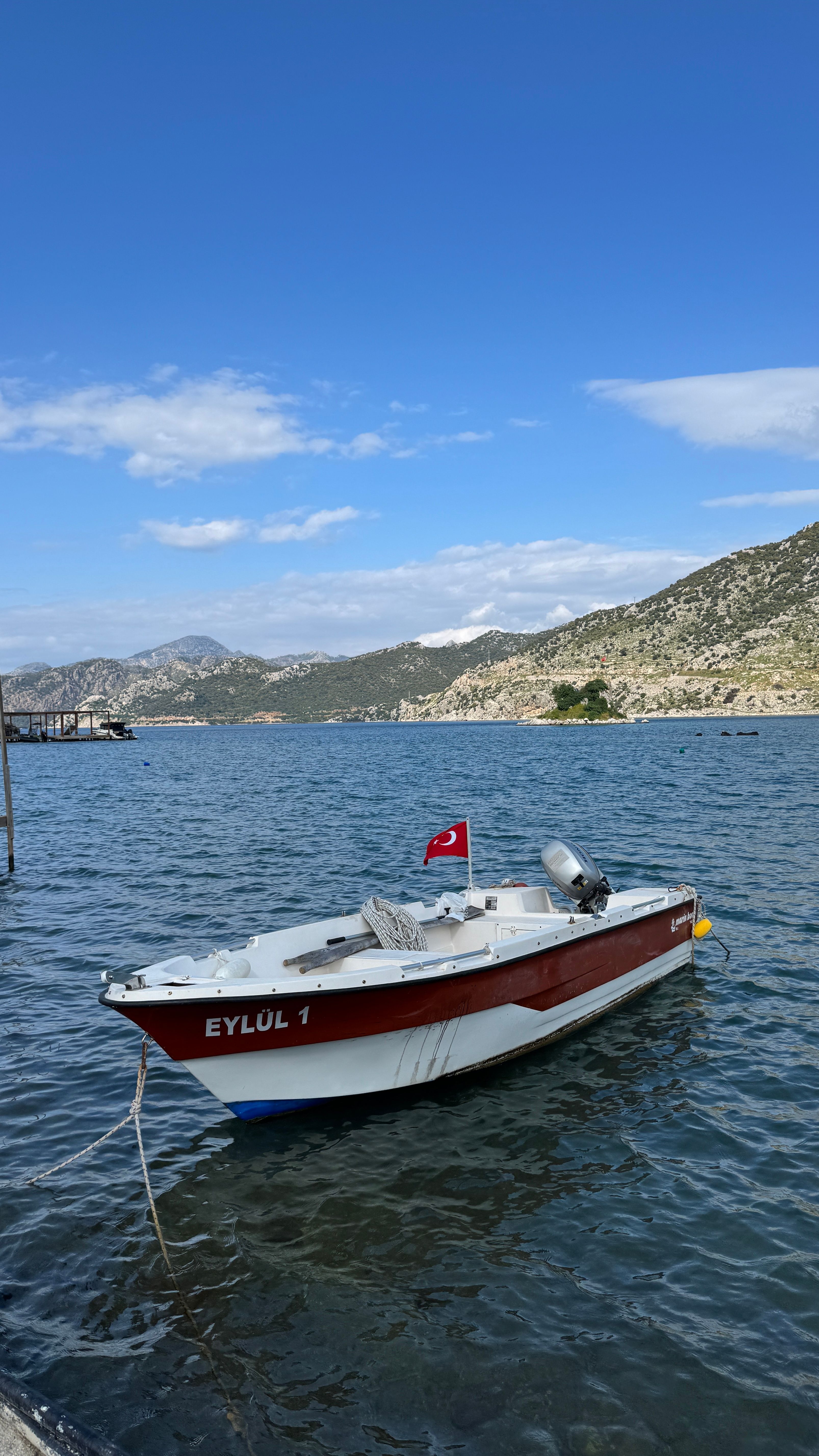 A boat named Eylol 1 with Turkish flag moored on the Aegean Sea, surrounded by mountains under a clear blue sky.