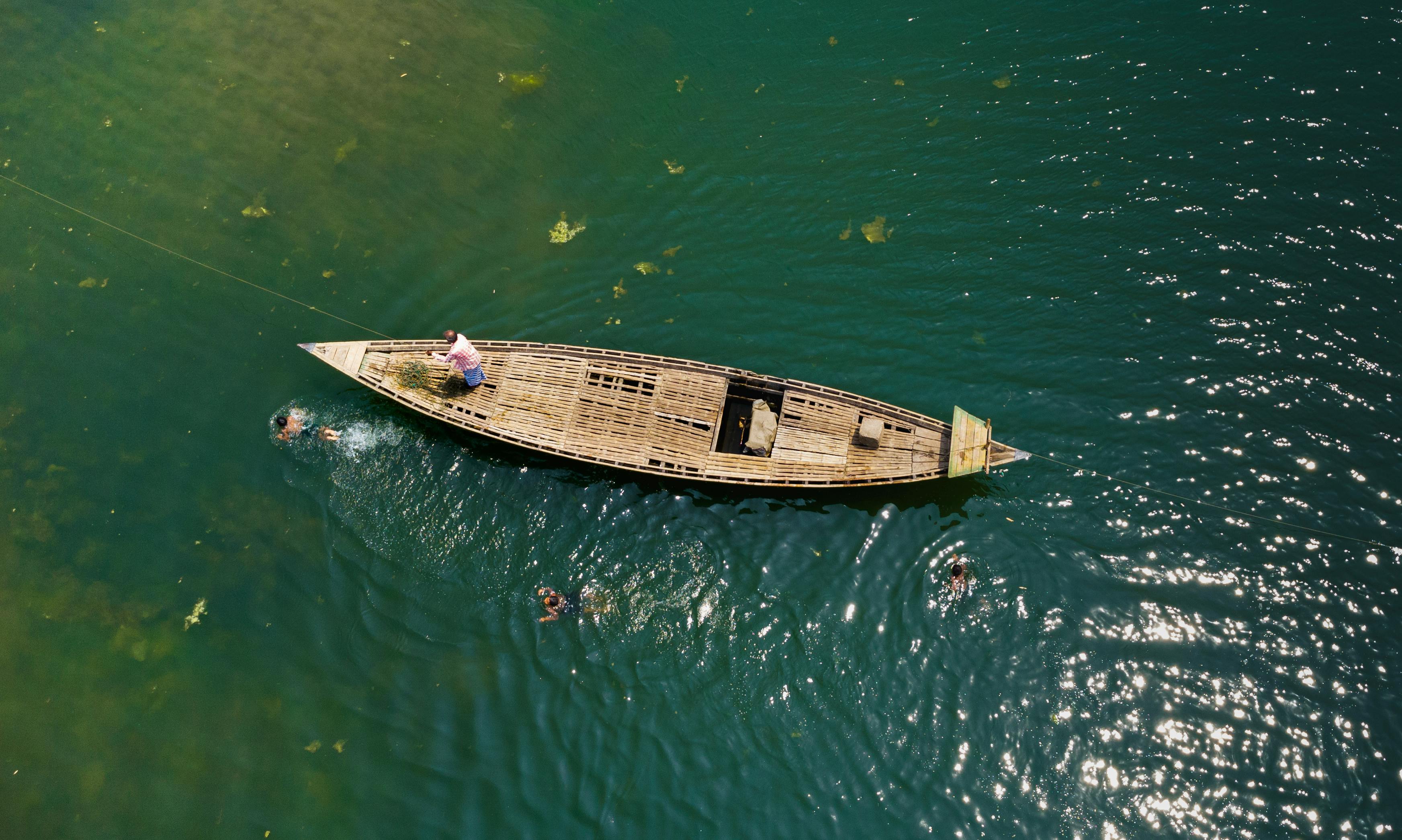 Aerial view of a traditional wooden boat with people swimming in a serene river.
