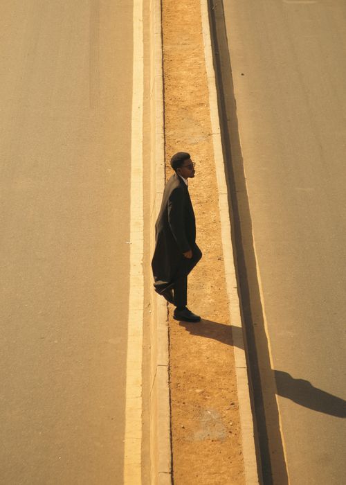 Free A solitary man in formal attire walks between divided road sections, casting a long shadow. Stock Photo
