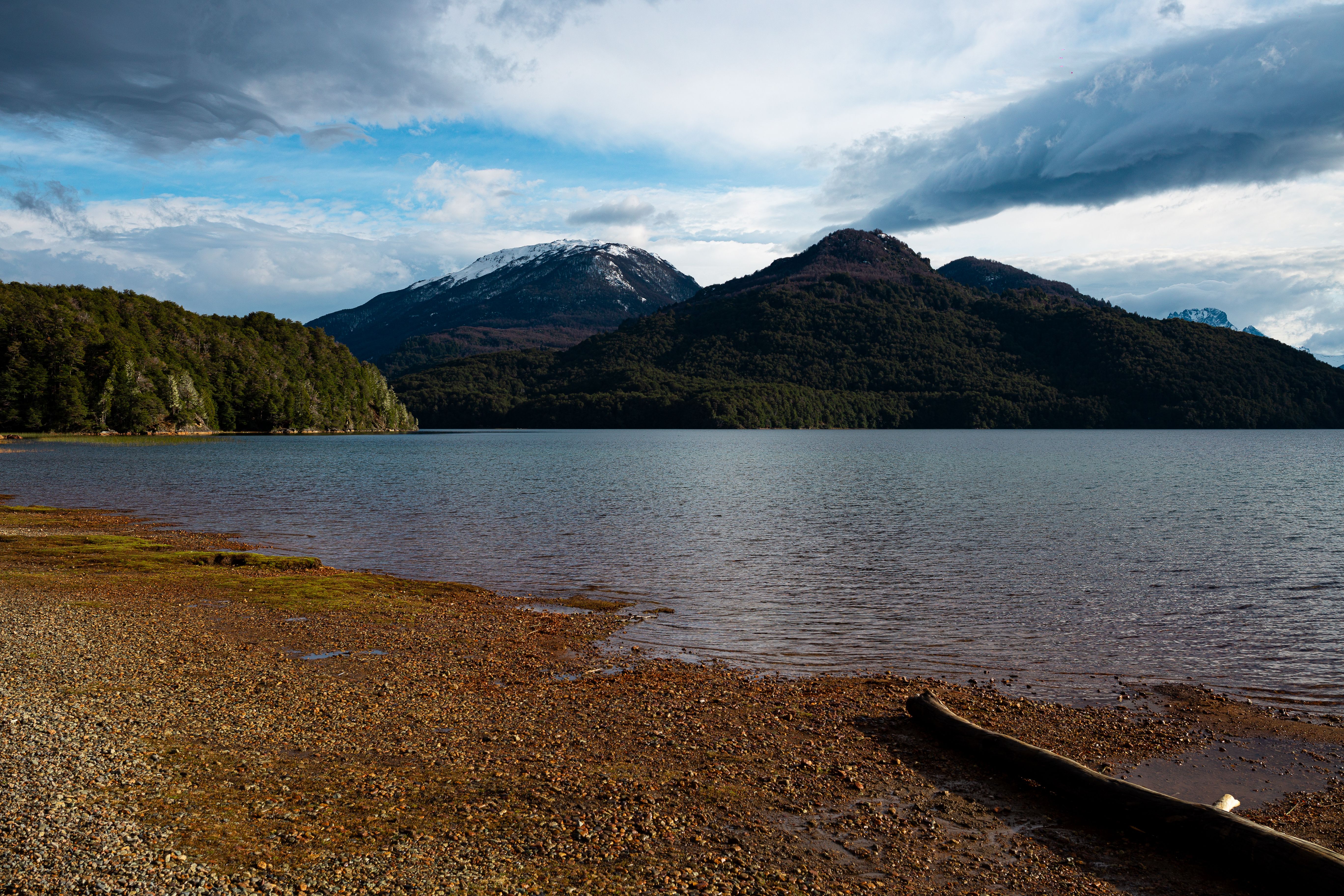 Tranquil lake with mountains, forest, and snowcapped peaks under a partly cloudy sky. Peaceful and scenic.