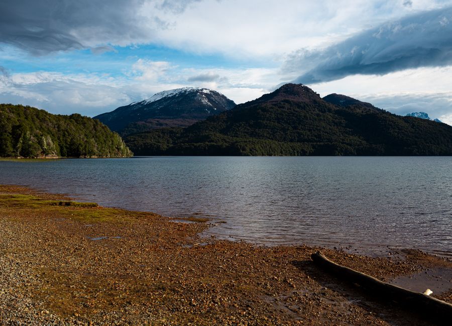 Tranquil lake with mountains, forest, and snowcapped peaks under a partly cloudy sky. Peaceful and scenic.