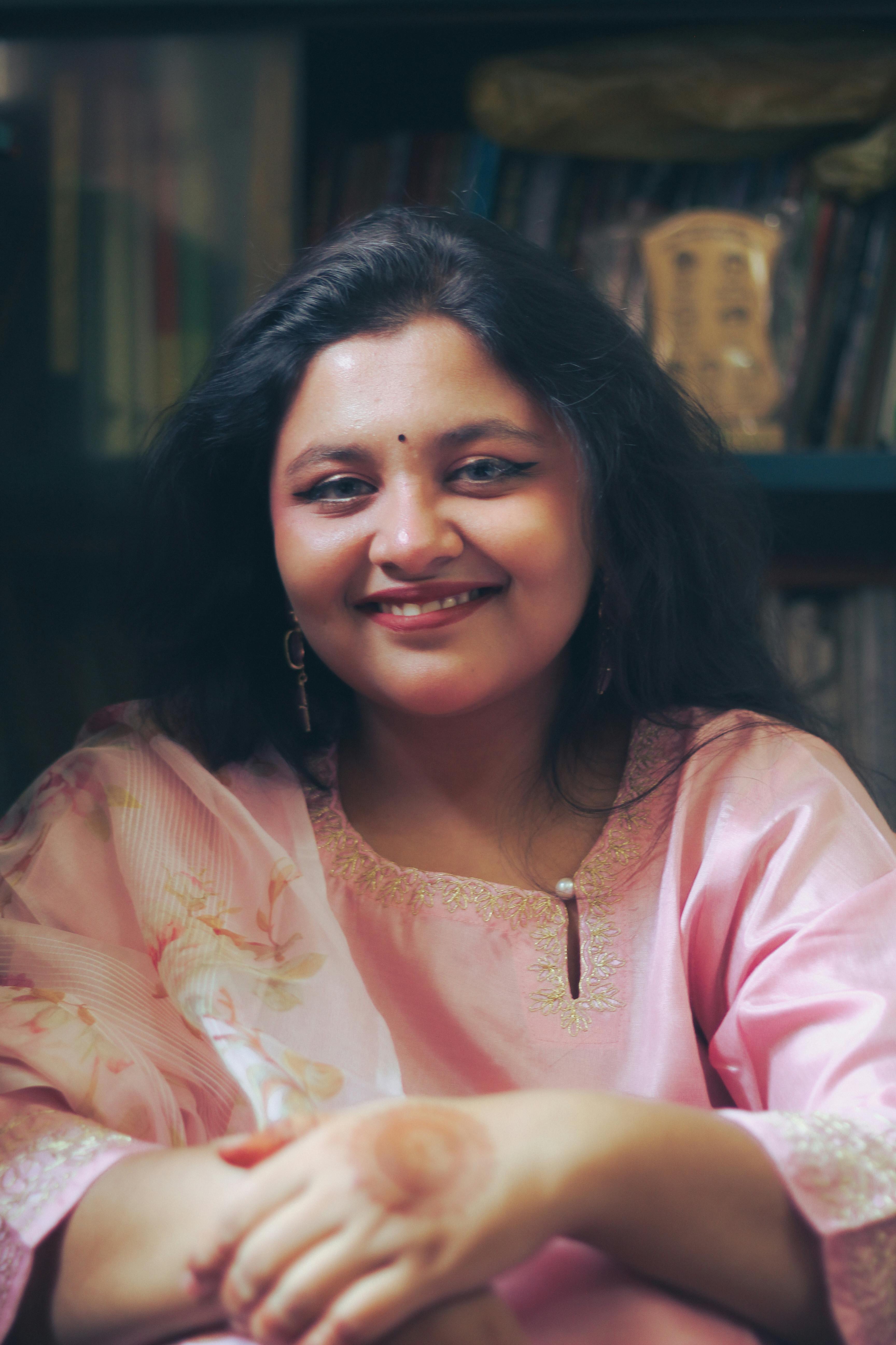 A woman in traditional outfit smiling warmly, seated indoors in Dhaka, Bangladesh.