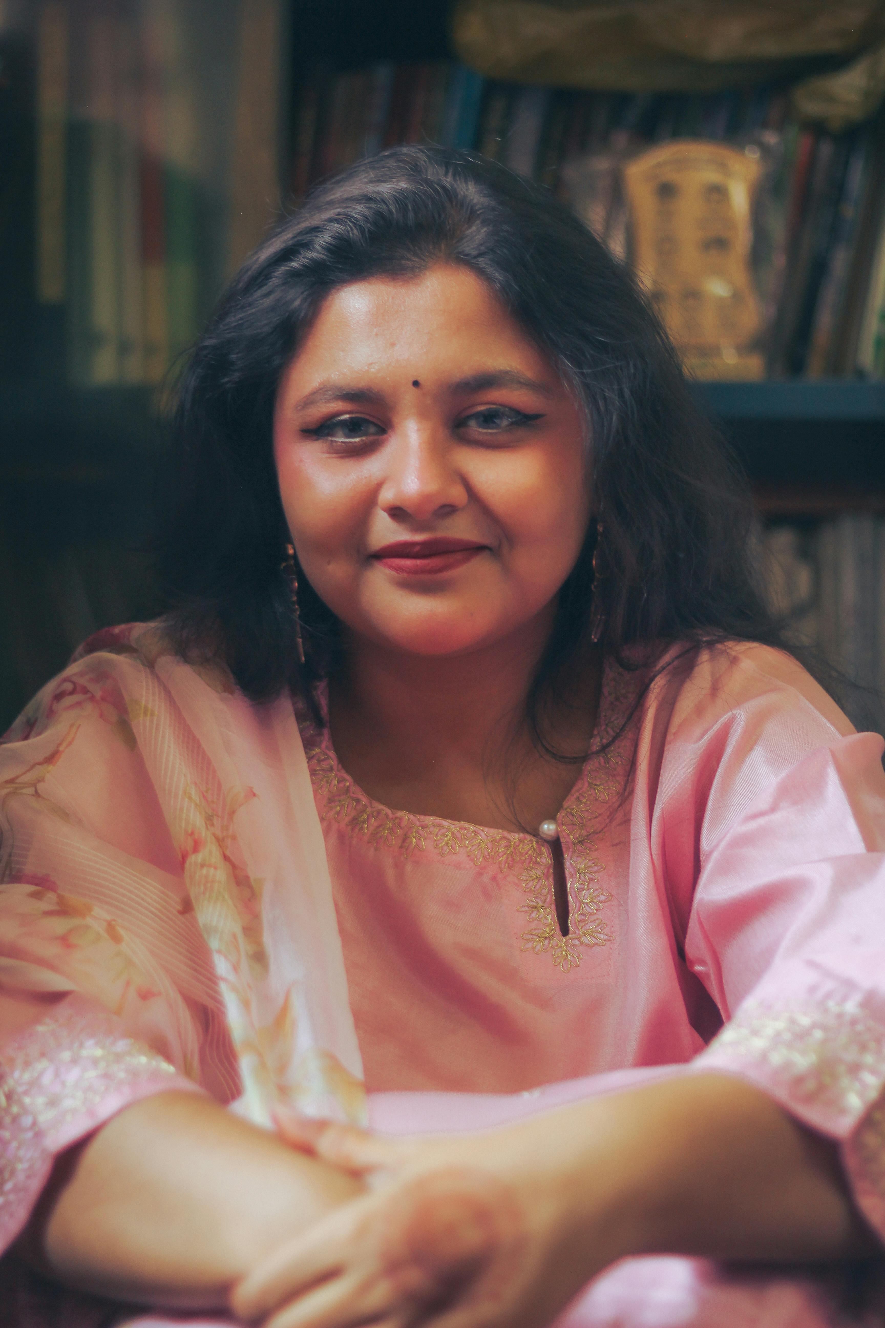 Portrait of a woman in a pink traditional dress, smiling indoors with books in the background.