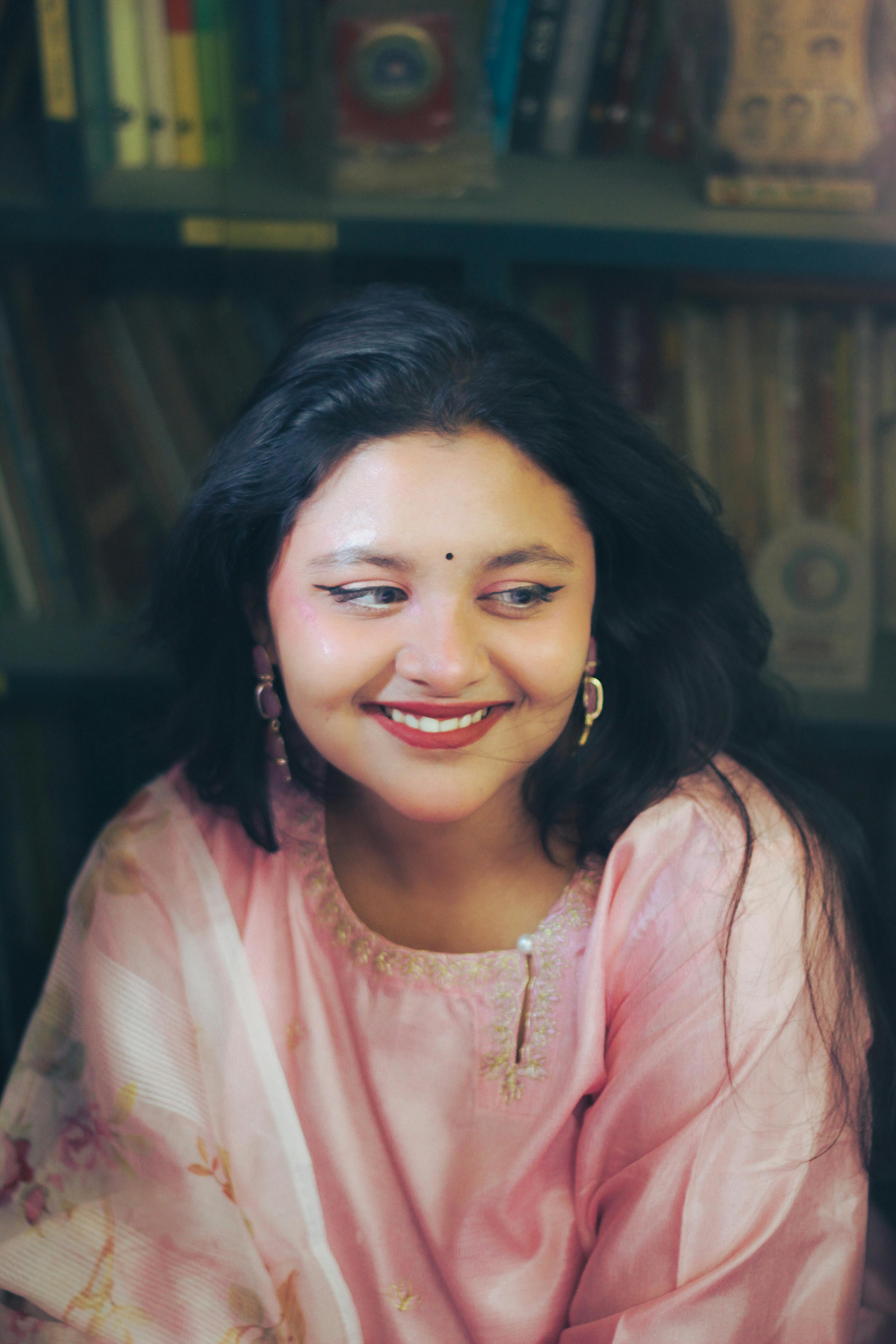 A South Asian woman smiling warmly in a pink traditional dress inside a library in Dhaka.