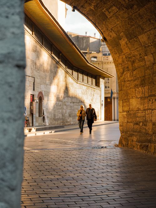 Free Two people walk through a sunlit street framed by an ancient archway, capturing the essence of a historical urban landscape. Stock Photo