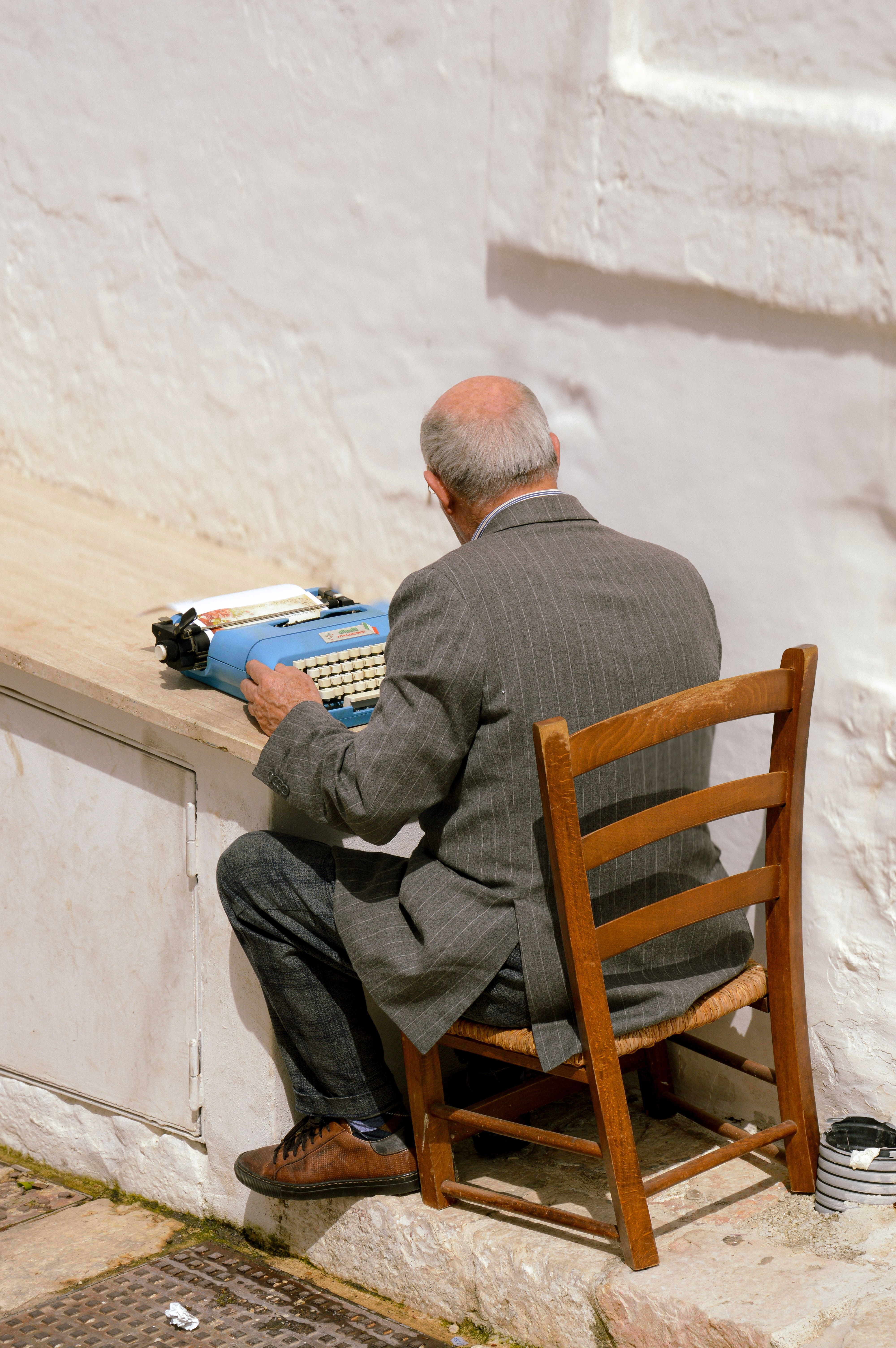 Elderly man using a typewriter outside in Alberobello, Italy, symbolizing nostalgia.