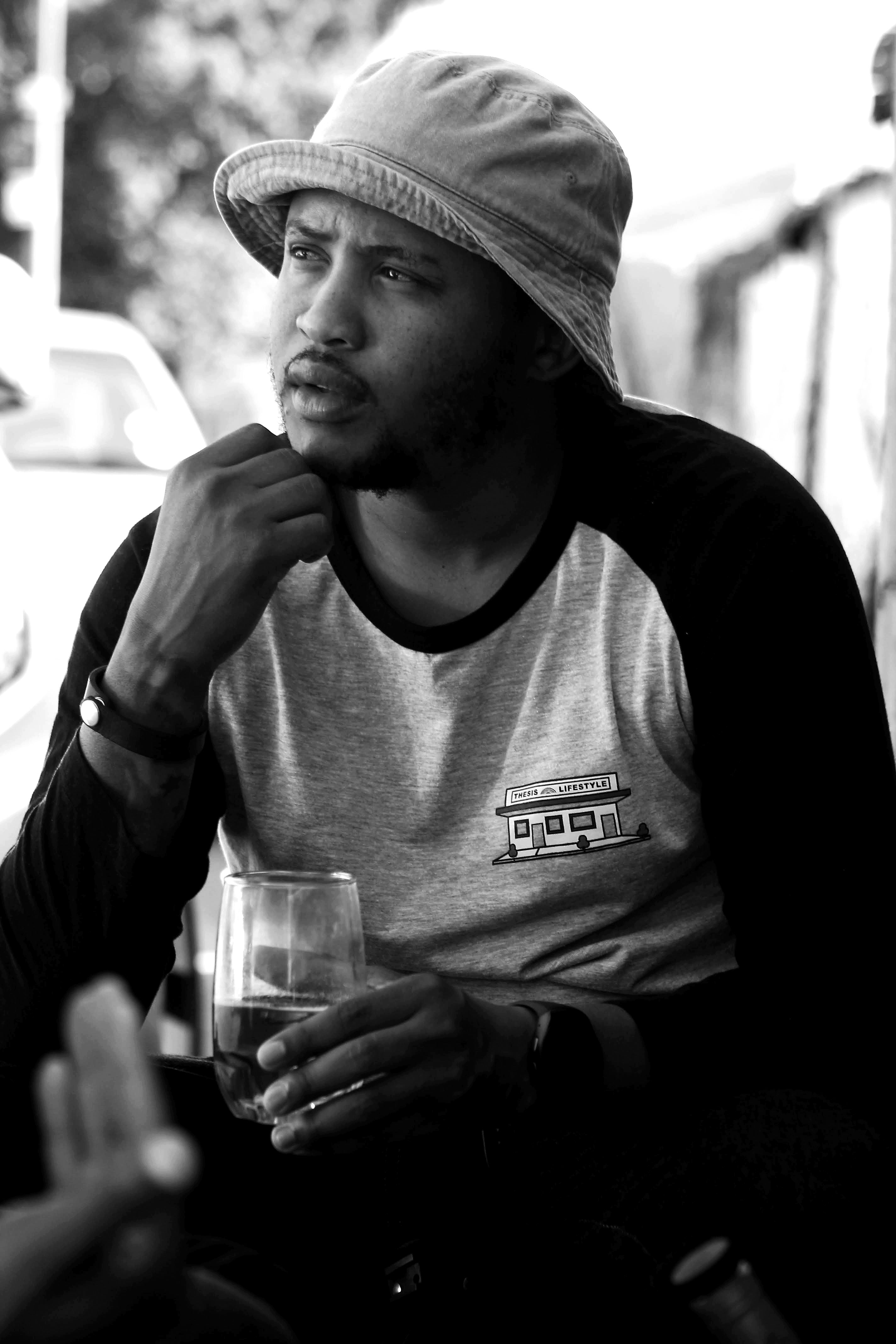 A man wearing a hat and casual attire holds a drink while sitting outdoors, lost in thought.