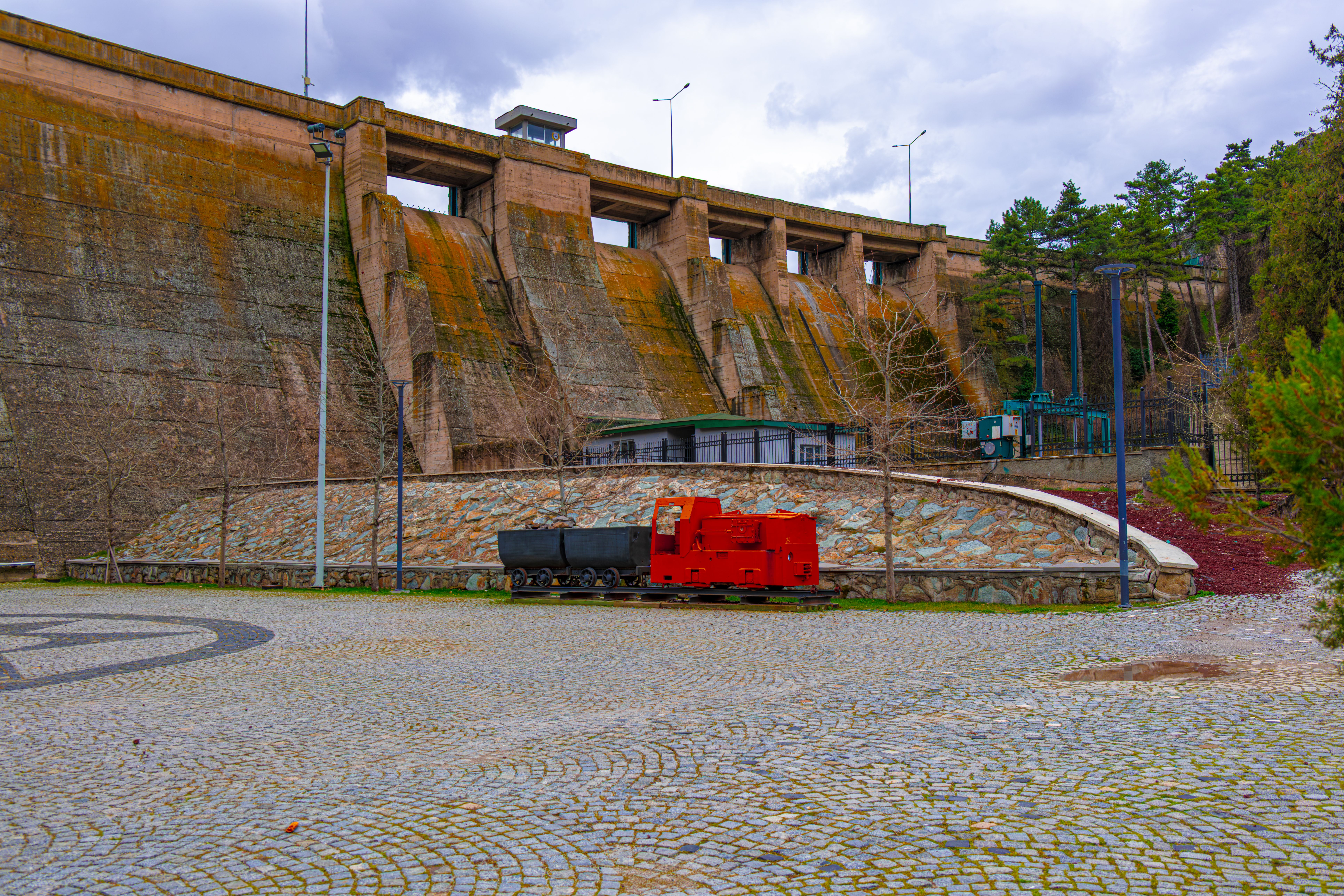 grátis Vista de uma antiga barragem em Ancara com uma locomotiva vermelha antiga sobre um pavimento de paralelepípedos. Foto profissional