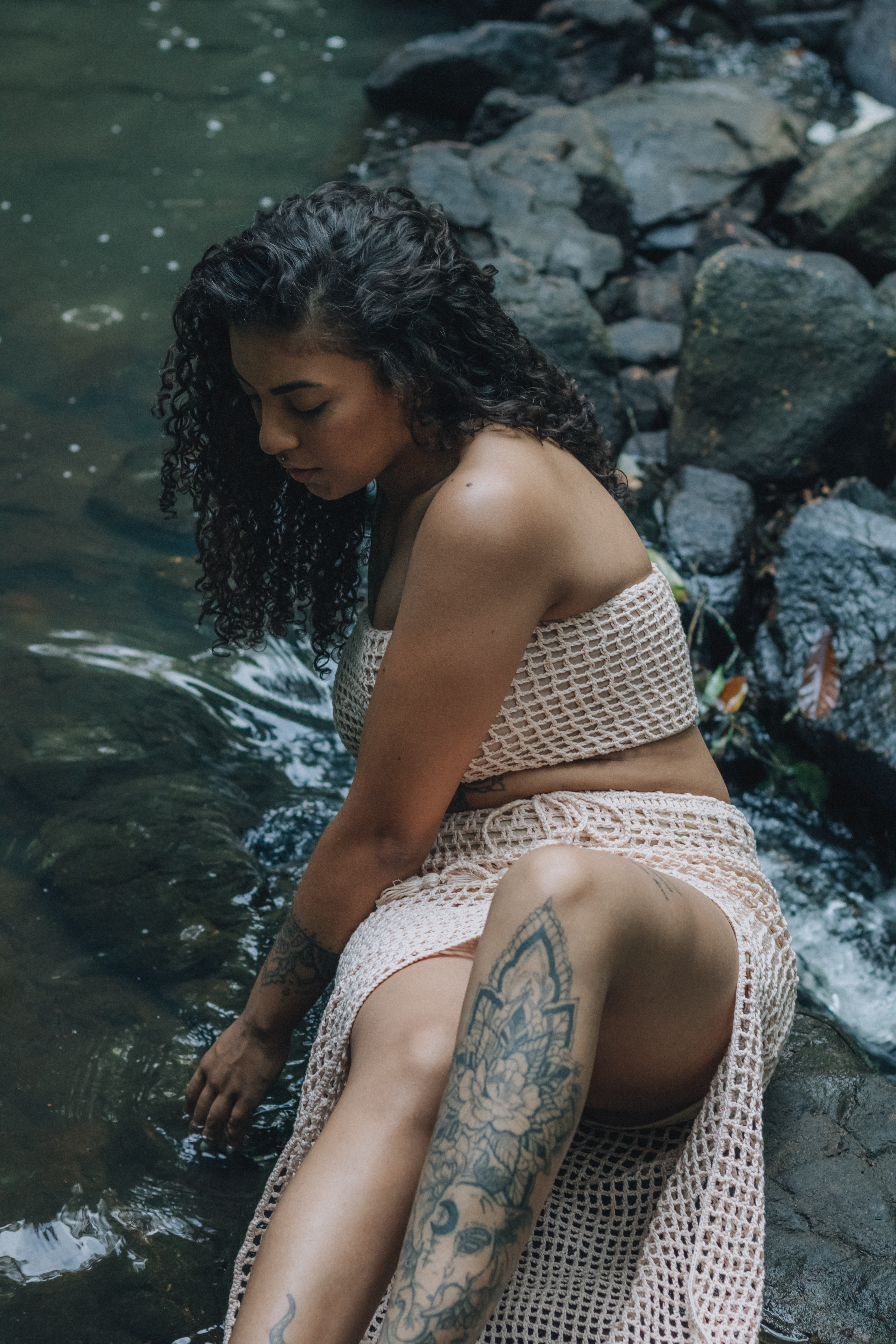 Woman with tattoo sitting by a rocky riverside, immersed in nature.