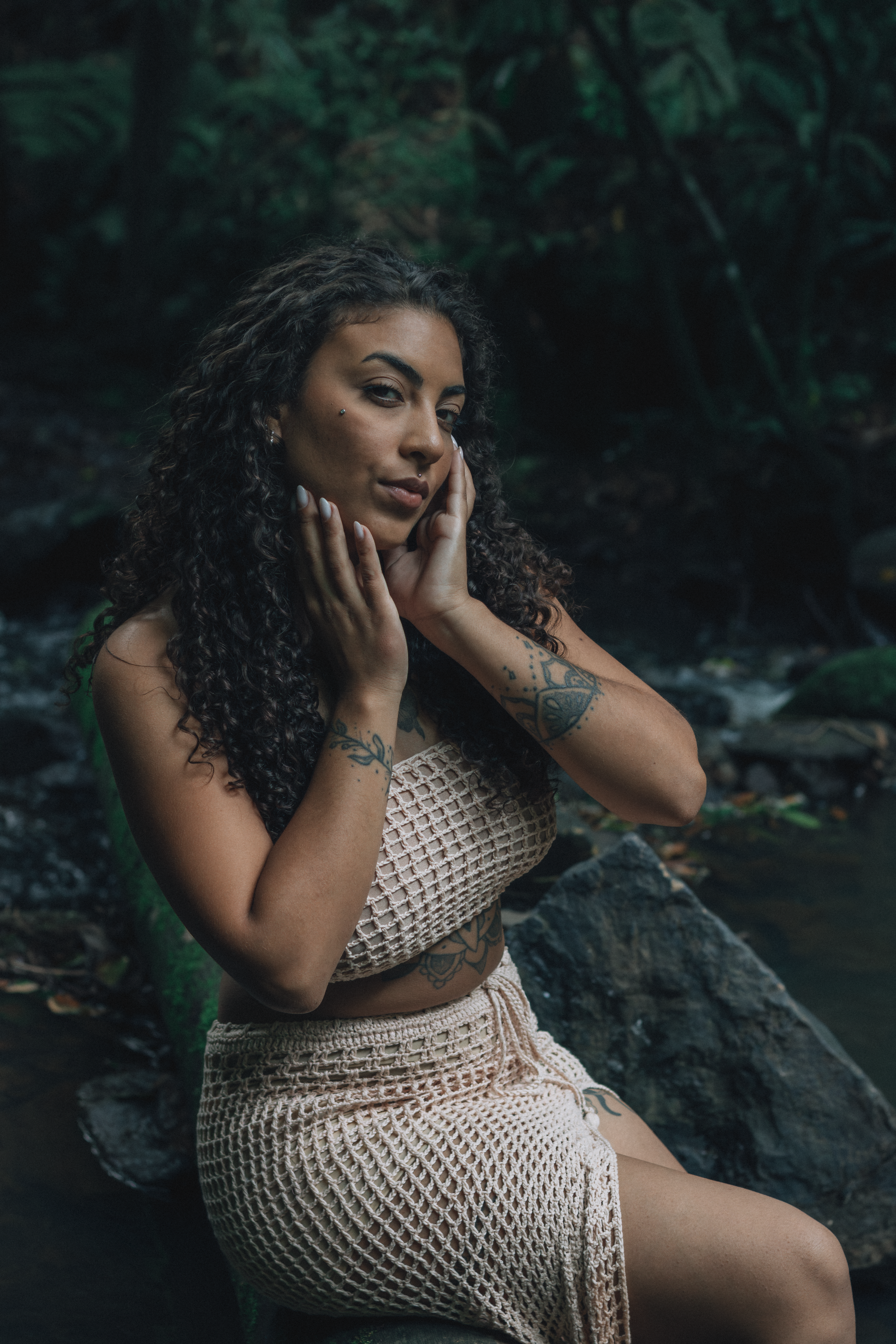 A young woman with curly hair poses thoughtfully by a tranquil stream in the forest.