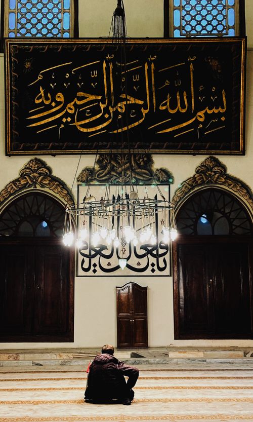 Free A man sits in prayer inside a historic mosque, under ornate Arabic calligraphy. Stock Photo