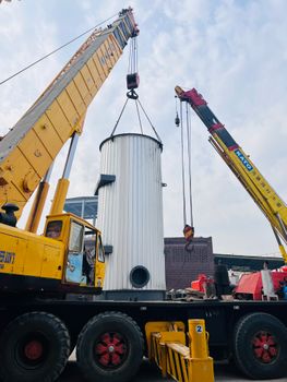 Two industrial cranes lifting a large white metal cylinder outdoors on a bright day.