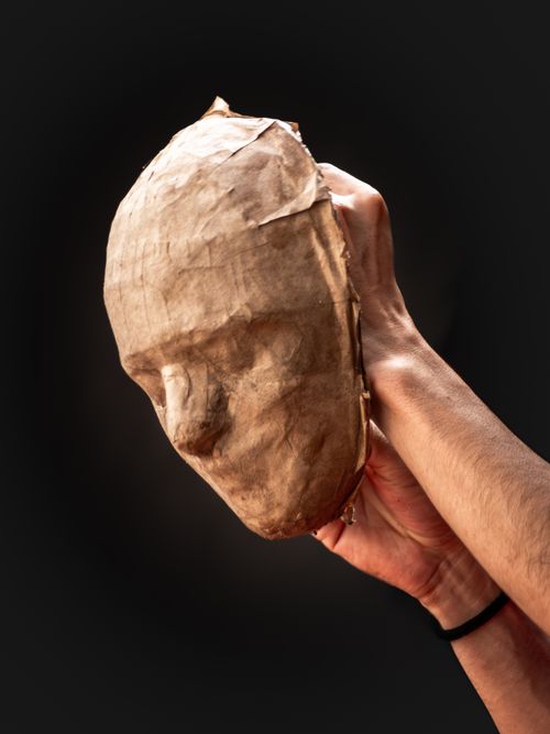 Free A hand holding a papier-mâché mask representing human features, set against a dark background. Stock Photo