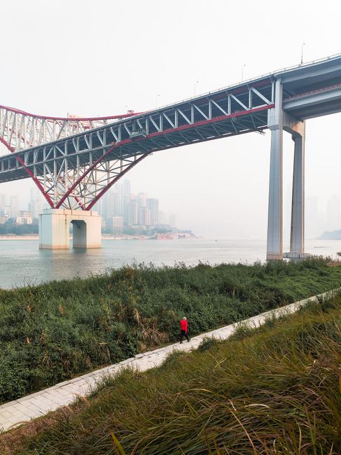 View of a large bridge spanning a river in a city with a person walking along a riverside path.