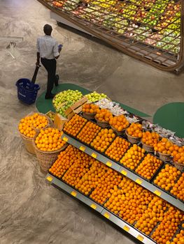 Aerial view of a vibrant fruit display with oranges and apples, as a shopper carries a basket in a supermarket aisle.