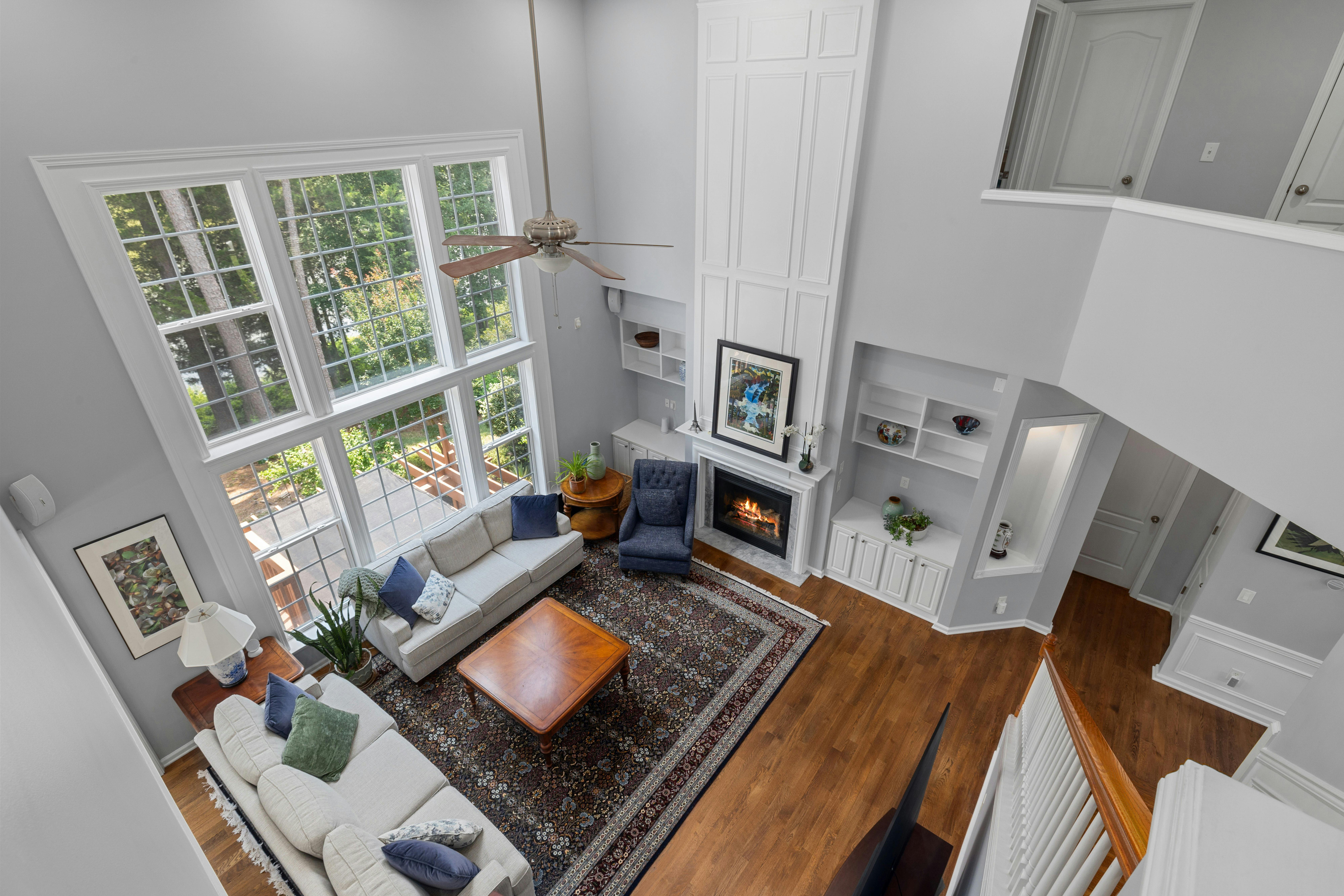 An elegant, spacious living room with soaring ceilings, large grid windows overlooking trees, a cozy fireplace, and classic furniture in warm and neutral tones.