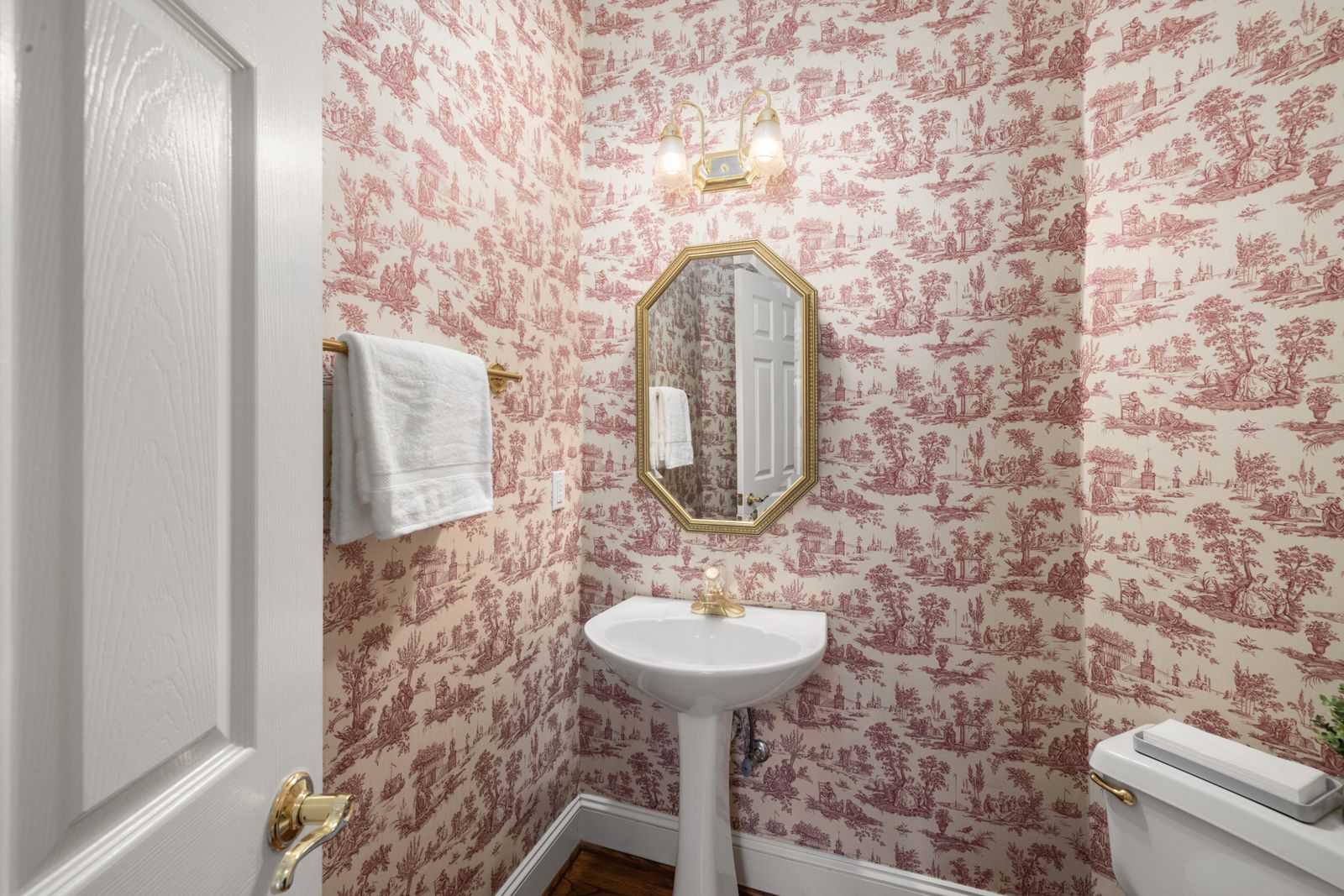 Red toile-style illustrated wallpaper around a pedestal basin and octagonal mirror.