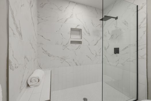 Elegant white marble shower with glass door, bench, and towel in modern bathroom.