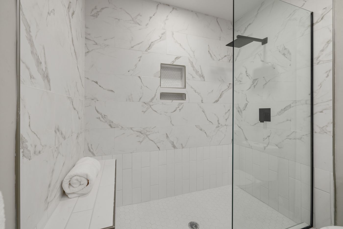 Walk-in shower with white marble-look wall tiles, a built-in niche, black shower fittings, frameless glass and small hexagonal floor tiles.