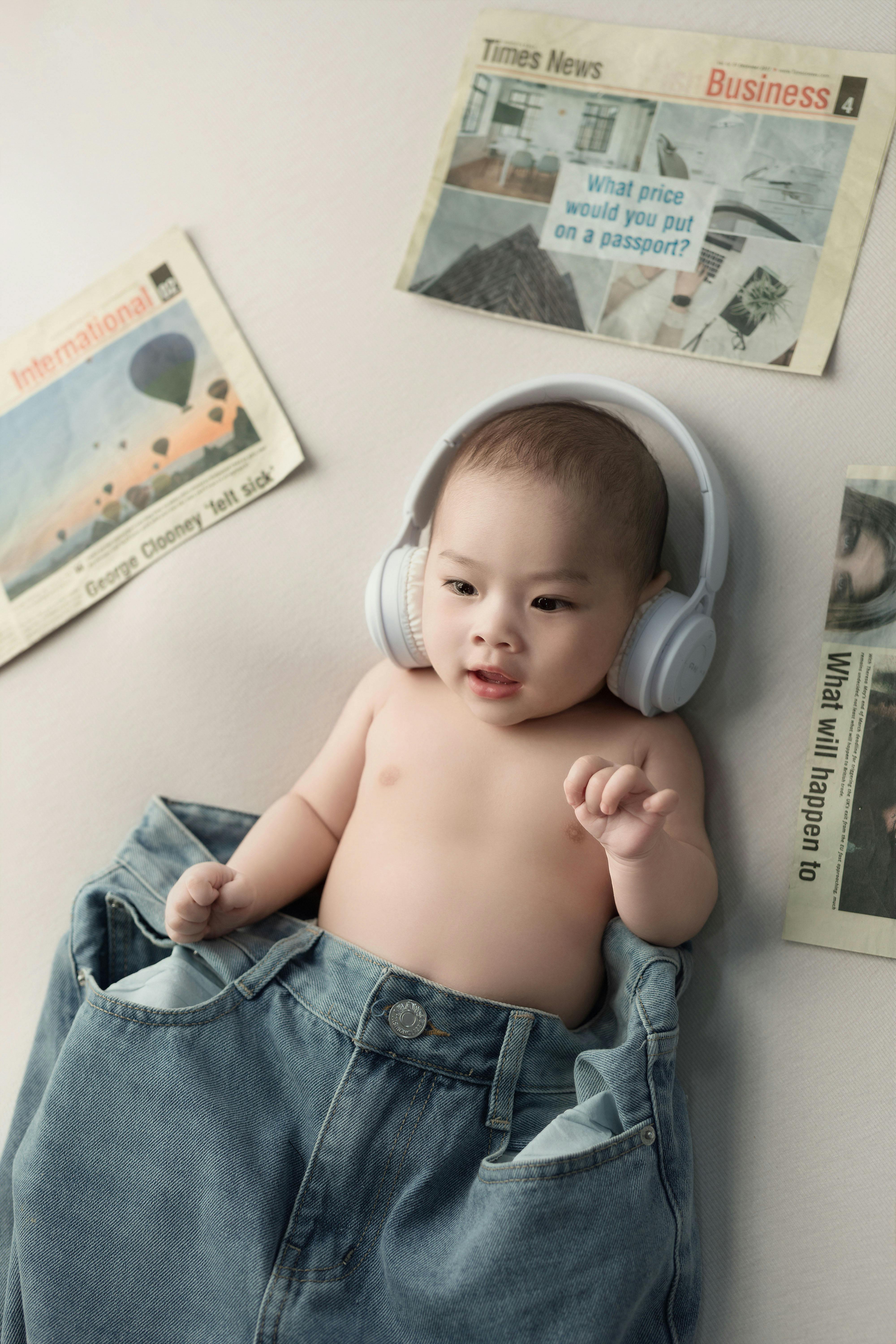 Cute baby wearing headphones and denim, surrounded by newspapers indoors. Modern lifestyle concept.