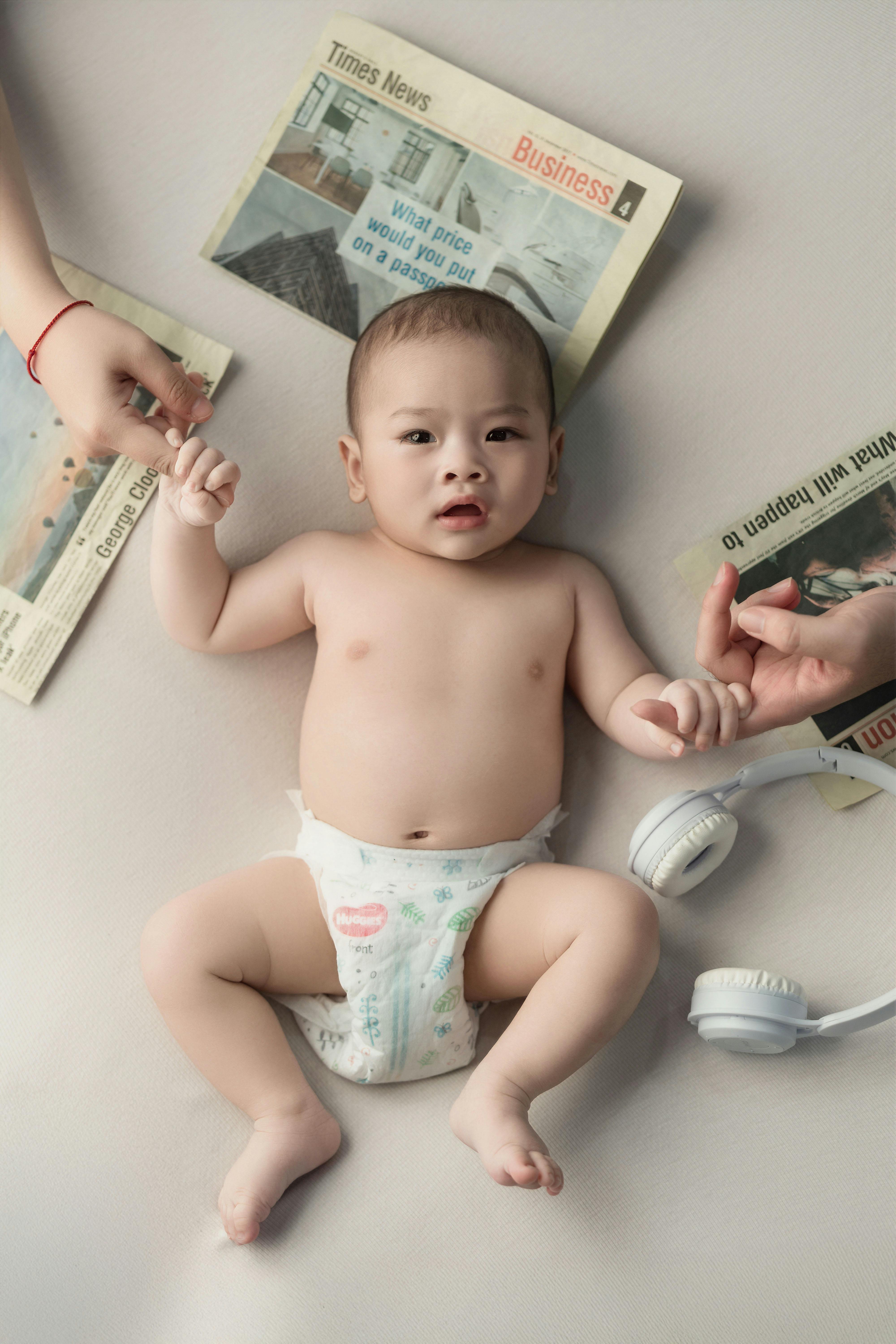 Cute baby laying on a bed surrounded by newspapers and headphones, holding parent's hand.