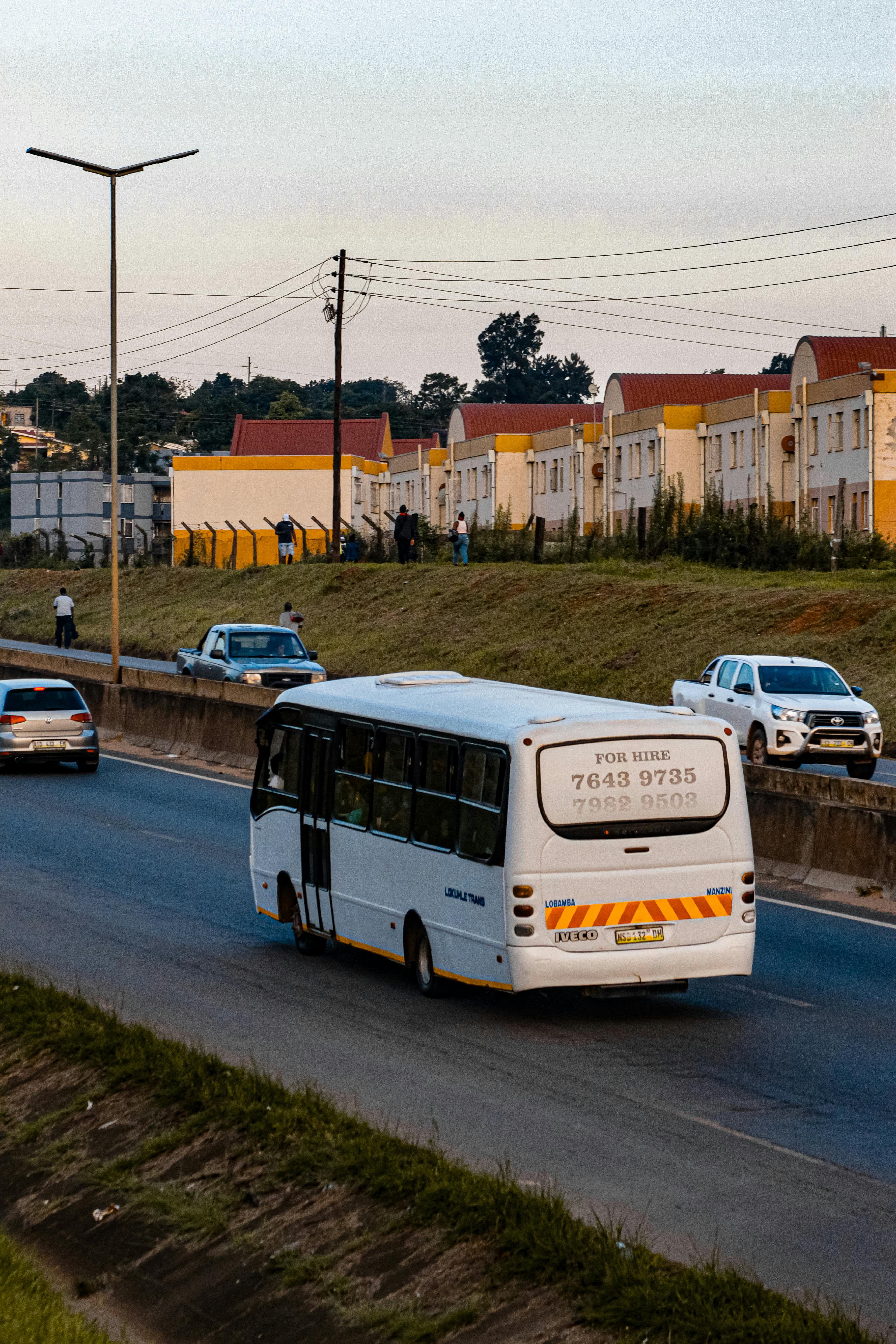Buses and cars travel on a highway in Matsapha, Eswatini near residential buildings during sunset.