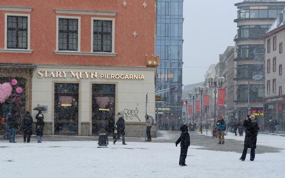 Winter snowfall at Stary Młyn restaurant corner, urban street view with pedestrians.