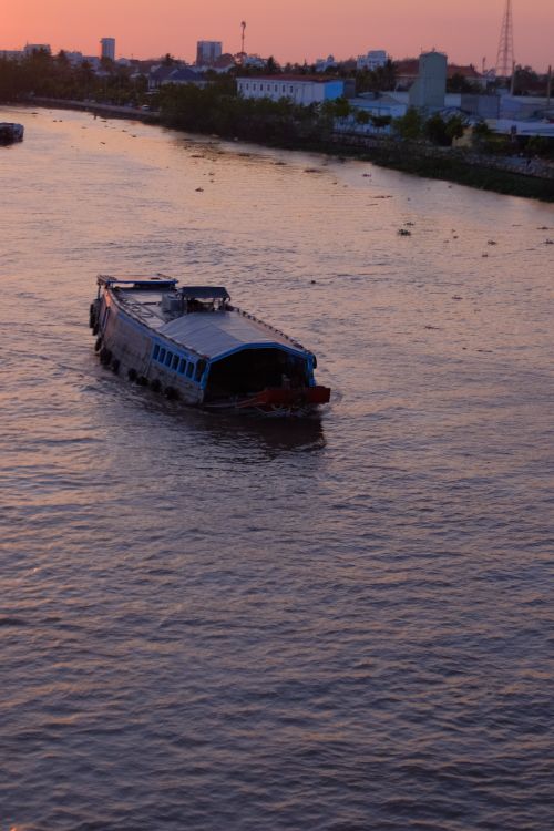 Free Boat cruising on a tranquil river during sunset with a city skyline in the background. Stock Photo