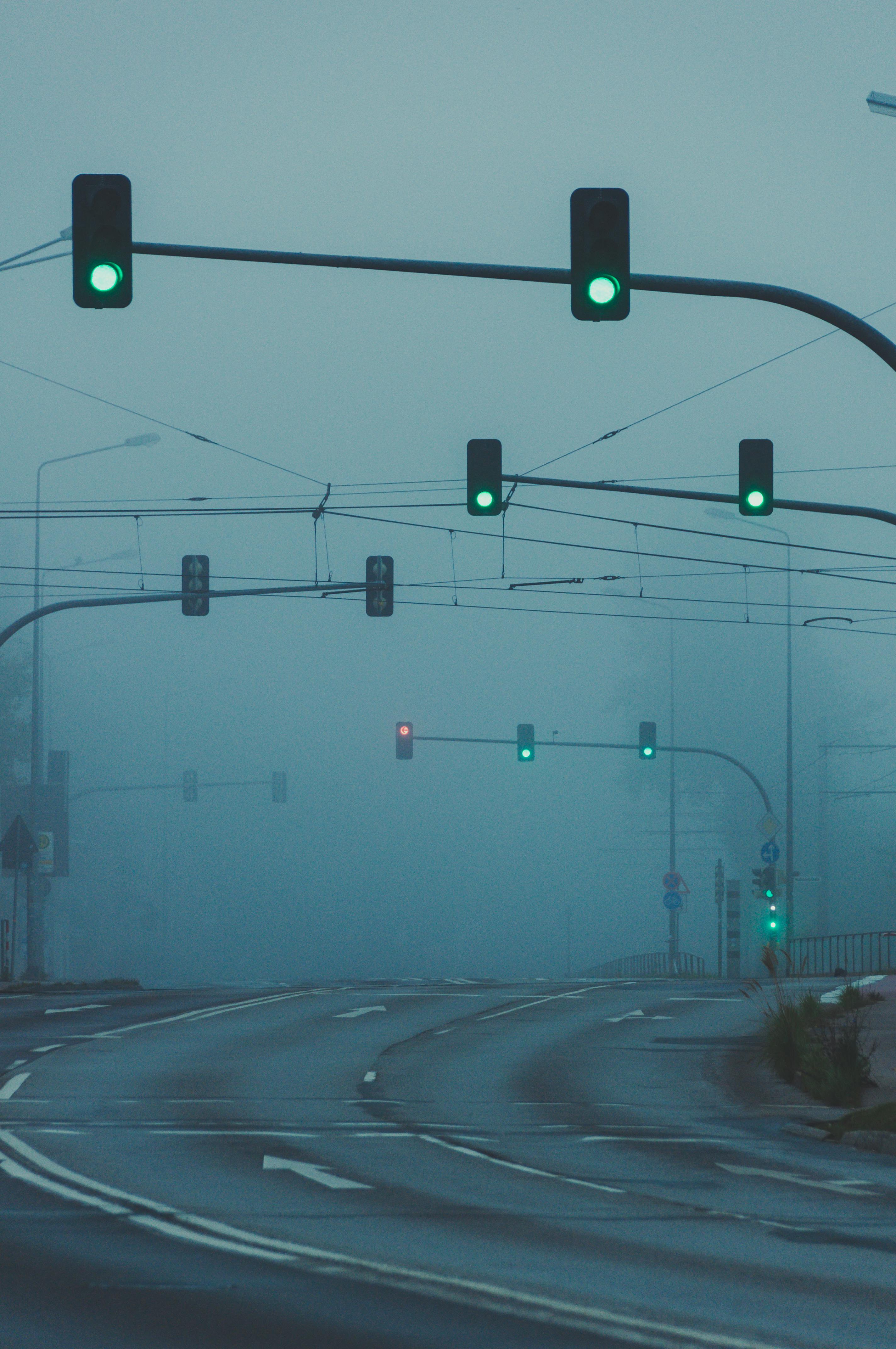 A fog-covered urban road intersection with multiple traffic lights glowing green.