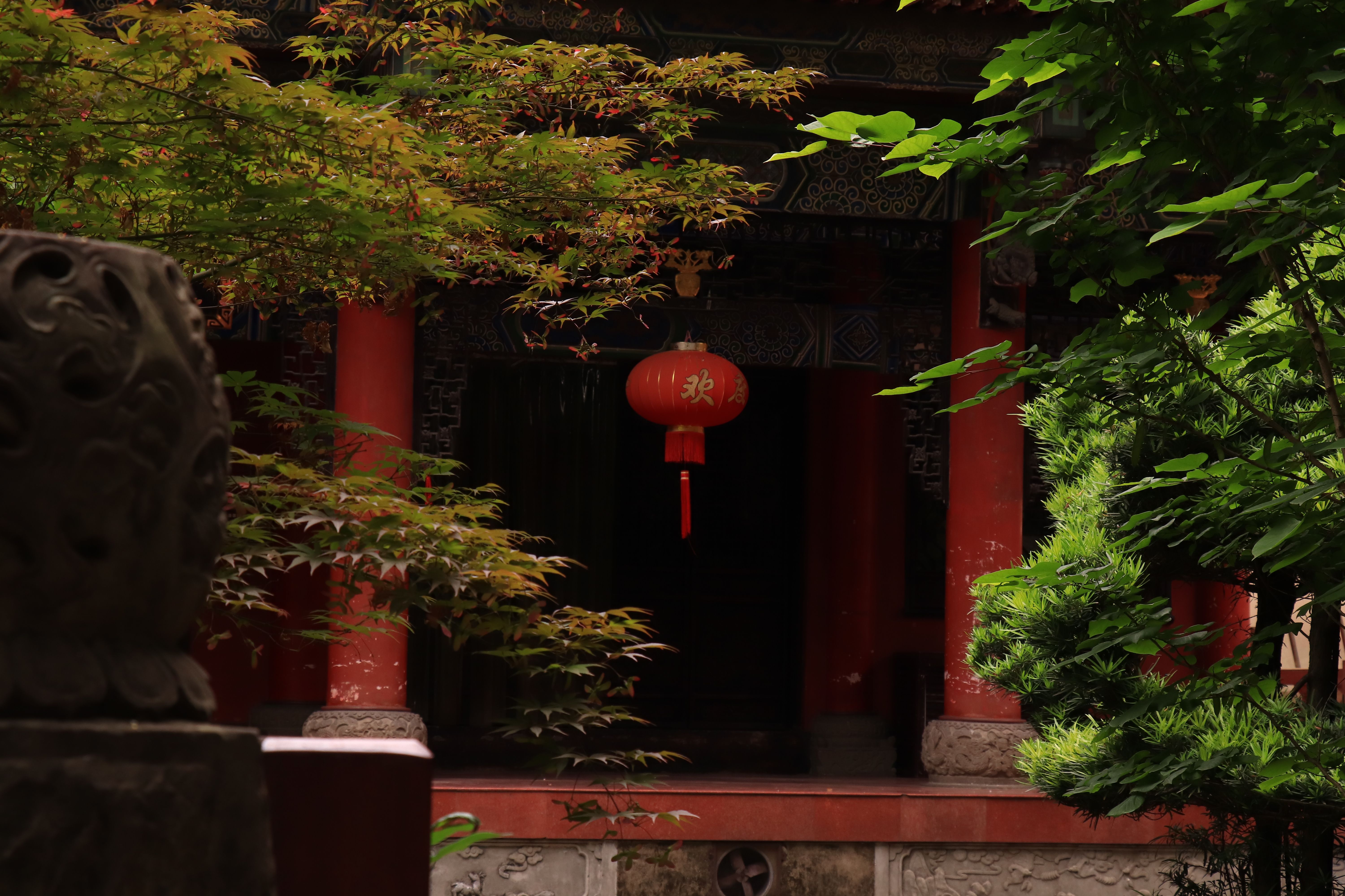 Ancient Chinese temple with red columns and lantern amidst lush greenery.