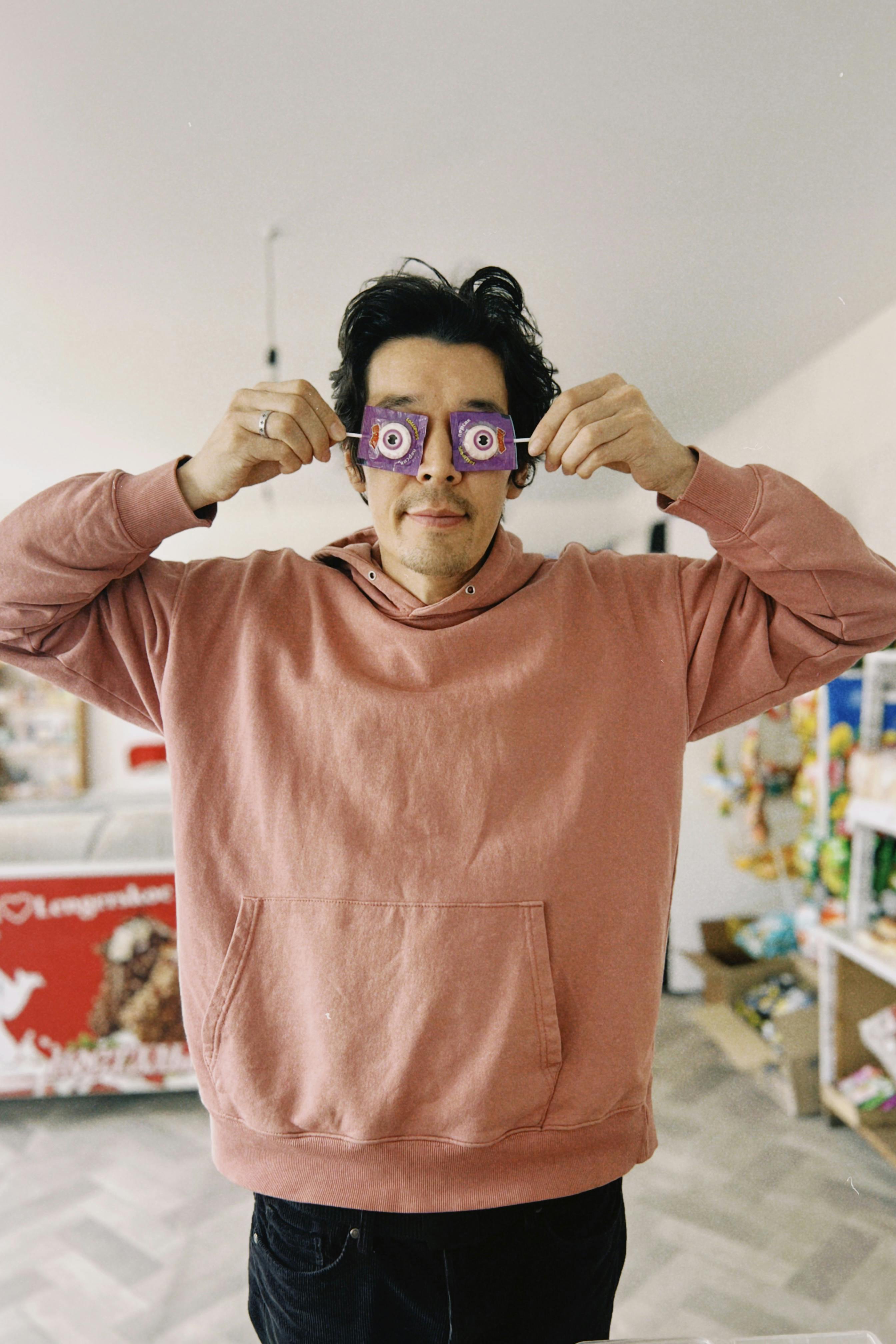 A playful man indoors, holding candy glasses in a humorous pose, wearing a pink hoodie.
