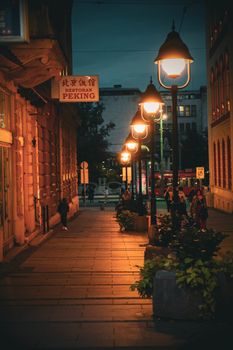 A moody evening street scene with glowing lamp posts and pedestrians in an urban environment.